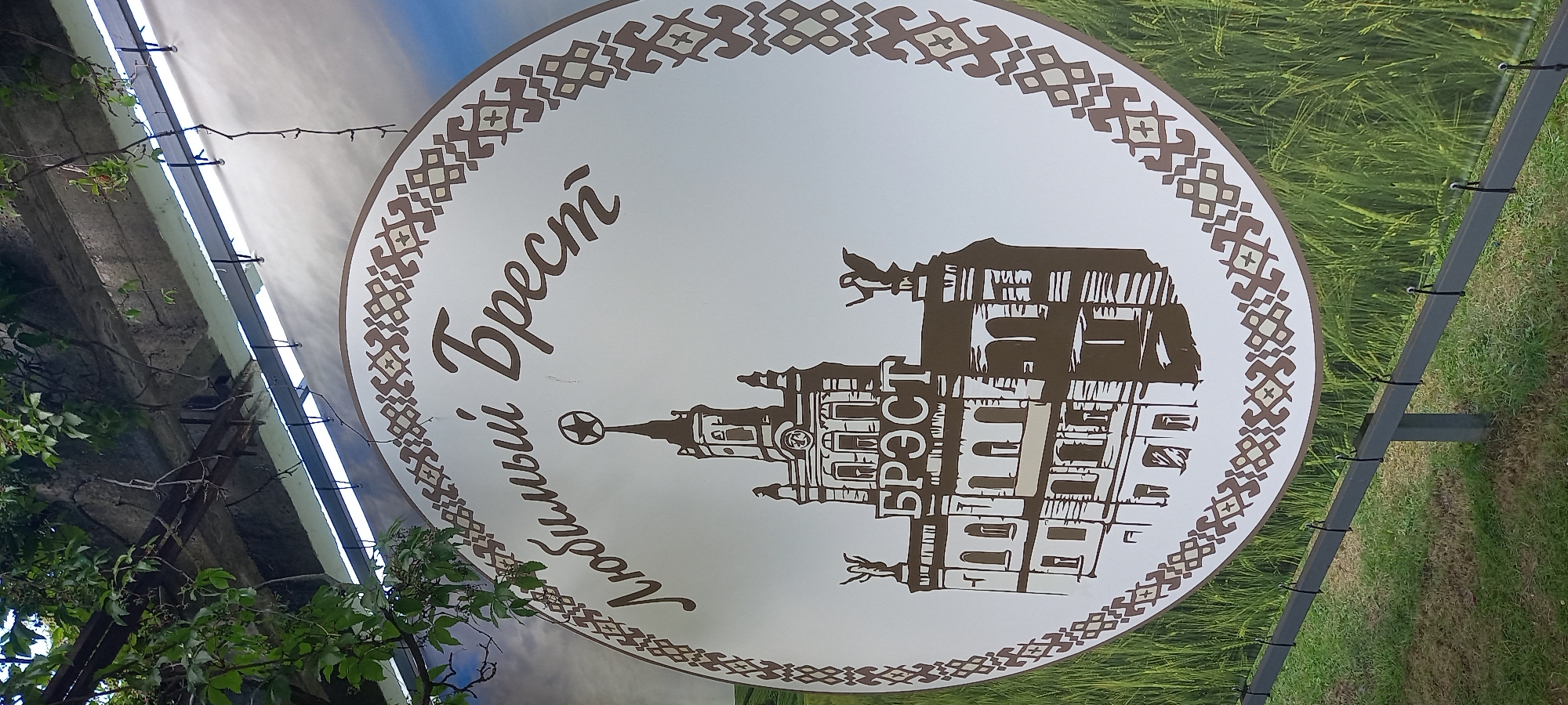 The image shows a circular sign with a stylized drawing of a building and text in Russian. The building is depicted in brown and white, with a star on top of its spire and two statues on either side. The text above the building reads "Любимый Брест" (Lyubimy Brest), which translates to "Beloved Brest." Below the building, the word "БРЕСТ" (BREST) is written. The sign has an ornate border with a repeating geometric pattern. The sign is hung outdoors, with green foliage from a vine visible at the top. In the background, there is a blurred image of a field of green grass or wheat. The sign is supported by a metal frame that is partially visible at the bottom of the image. The overall scene suggests it is an outdoor advertisement or landmark sign for the city of Brest, Belarus.