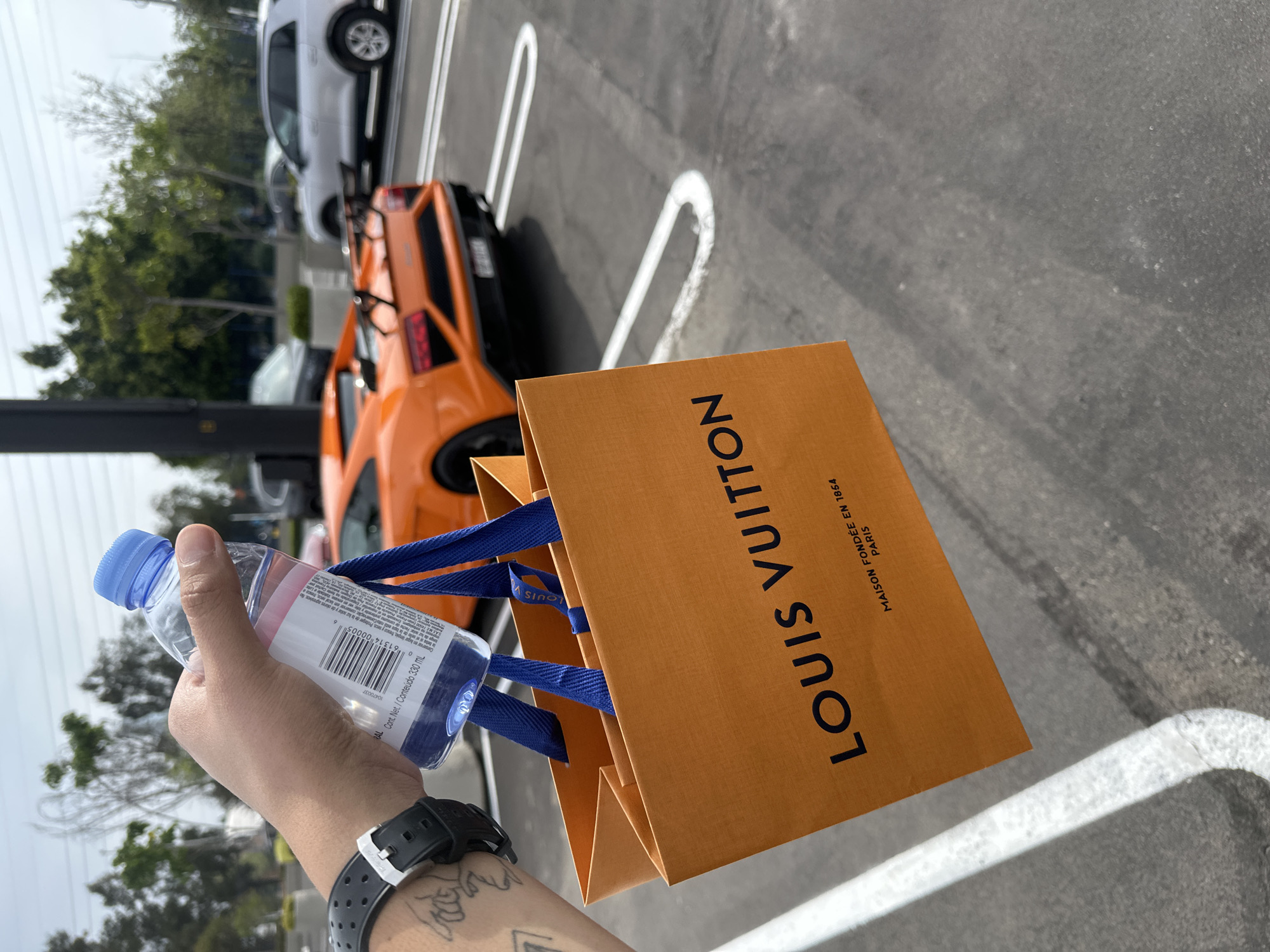 A man holds a water bottle and a Louis Vuitton shopping bag outdoors. In the background, there is a parked orange Lamborghini and a white vehicle, located on an asphalt surface marked with white parking lines. The scene is daytime and appears to be in an urban area. No information is provided about the location in Puebla, Mexico.