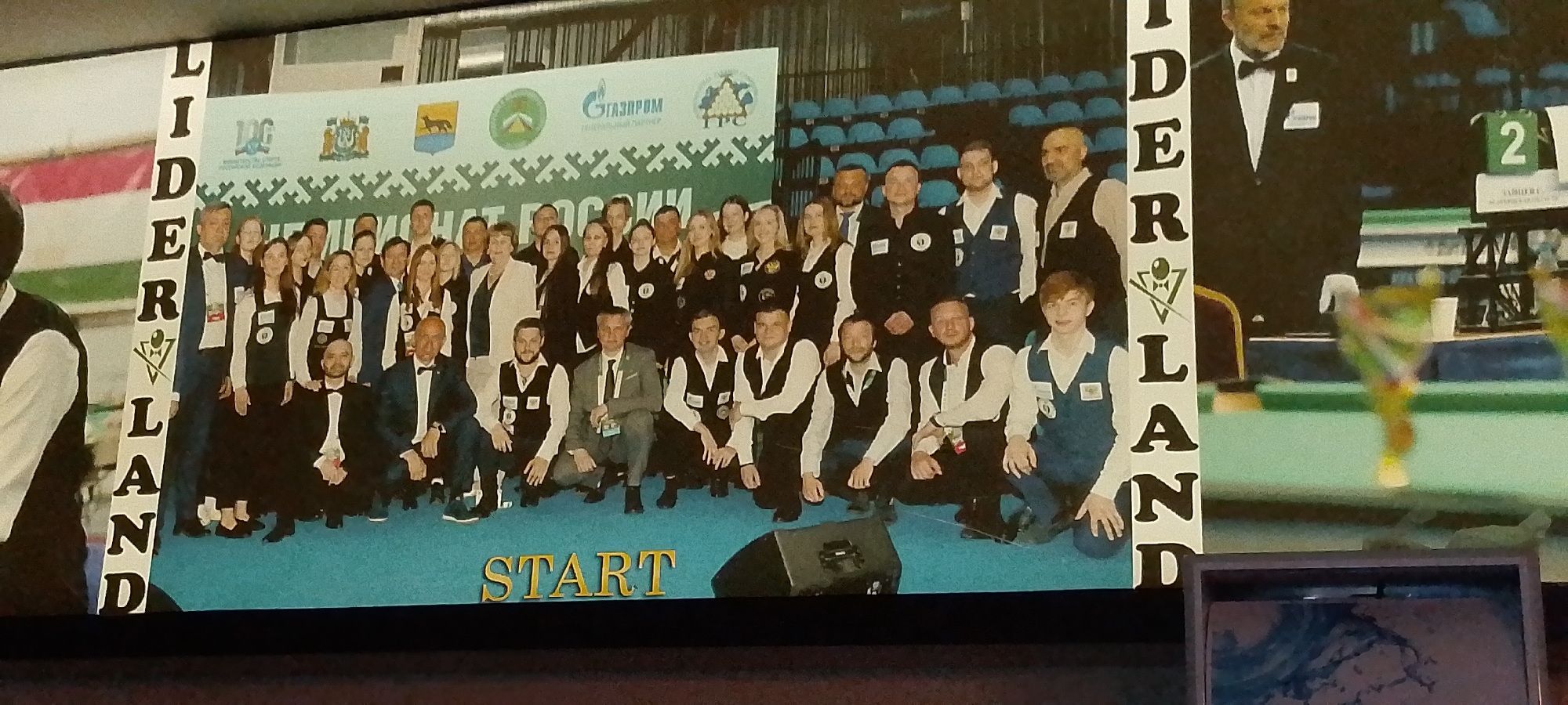 The image shows a large group of people, a mix of men and women, posing for a photograph. They are dressed in formal attire, mostly black suits with white shirts and bow ties, and some are wearing vests. In the background, there is a large banner with text and logos, including "ГАЗПРОМ" and "ТПС". The word "START" is written in large golden letters on the floor in front of the group. Some of the individuals are kneeling, while others are standing. The environment appears to be an indoor event, possibly a sporting competition or an awards ceremony, given the presence of what looks like a billiard table or similar green surface in the background and trophies on display. The phrase "LIDERLAND" is written vertically on the left side of the image, with a stylized logo incorporating a cue and balls. The overall impression is of a team or a delegation being photographed. The image does not contain any information that indicates the location is in the city of Angren, Uzbekistan.