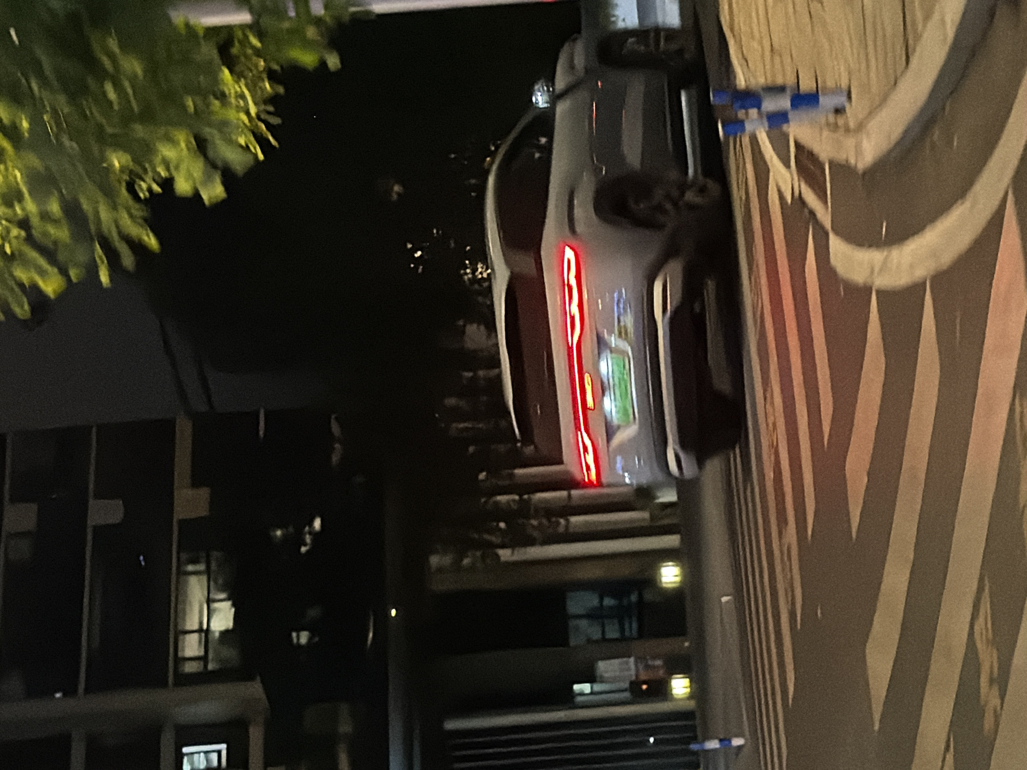 In the city of Jiangjin, China, at night, a light-colored SUV is captured in motion on a street at a crosswalk. The vehicle, seen from the rear, displays illuminated red taillights and a green license plate. The surroundings include a modern building with illuminated windows, a tree with visible green foliage in the upper right, and a sidewalk with blue and white bollards. The crosswalk stripes are clearly visible on the asphalt road.