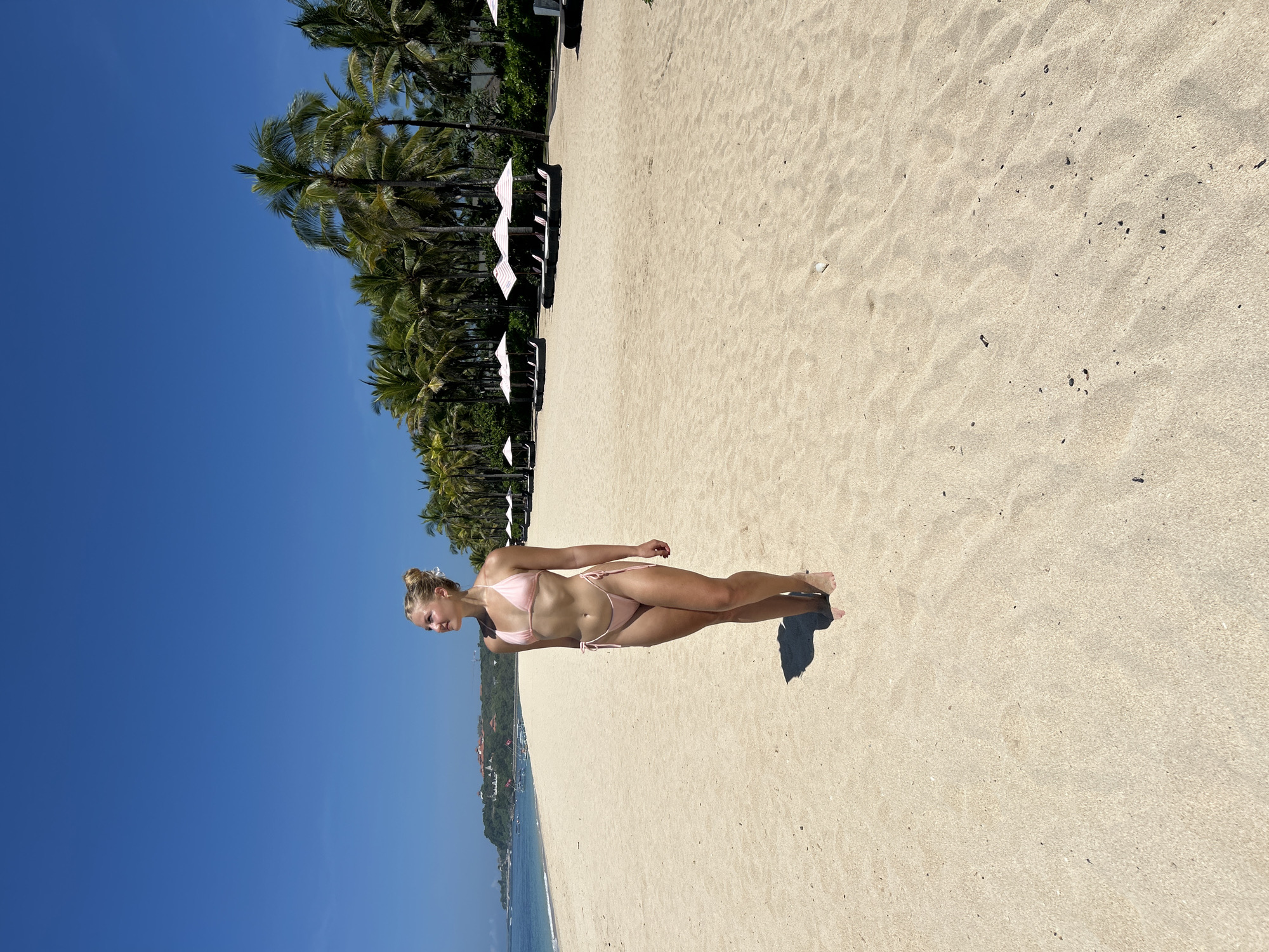 A woman in a pink bikini stands on the sand in Kuta Selatan, Indonesia. She is facing left. The beach is sandy. Palm trees and white umbrellas line the edge of the beach. The sky is clear blue. The water is visible in the distance.