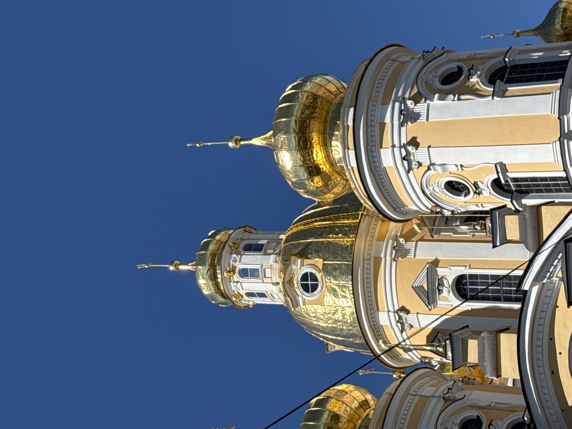 The image shows the gilded domes and yellow and white facade of a church against a clear blue sky. The church features multiple domes of varying sizes, topped with golden crosses. The architecture is ornate, with decorative moldings, pilasters, and arched windows. The scene is captured from a low angle, emphasizing the grandeur of the religious structure. This appears to be a section of the Smolny Cathedral in Saint Petersburg, Russia, known for its distinctive Baroque architecture and blue and white exterior. There are no people or animals visible in the frame.