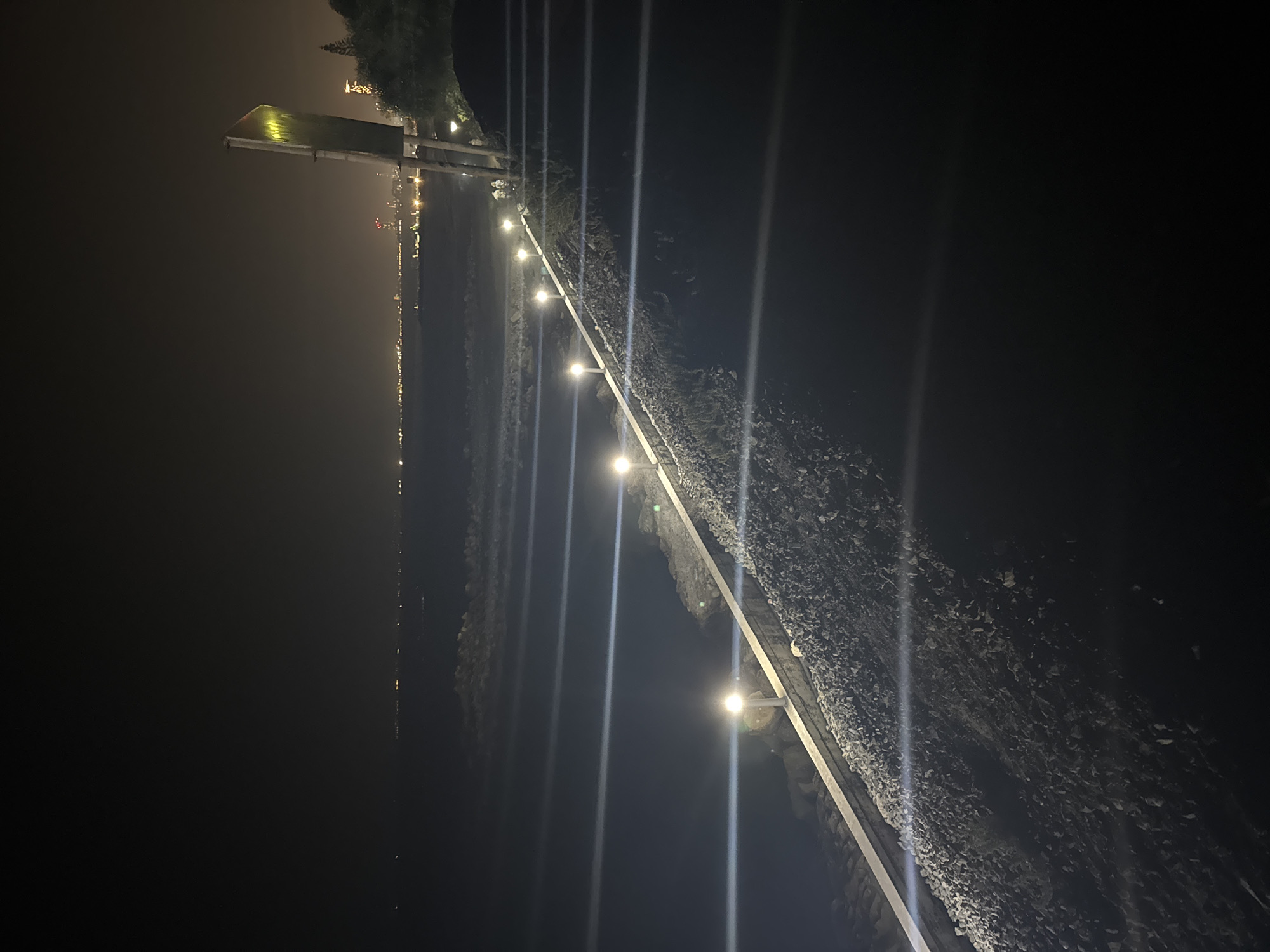 A long shot of a coastline at night shows a paved walkway with evenly spaced lampposts casting light on the gravel and water. The walkway is bordered by a white railing and a rocky embankment. In the distance, city lights twinkle along the horizon, suggesting a developed area. A signpost stands to the right, its surface reflecting the ambient light. The sea is calm, with gentle waves lapping the shore. The overall atmosphere is serene and tranquil. The location is Agios Tychonas, Cyprus.