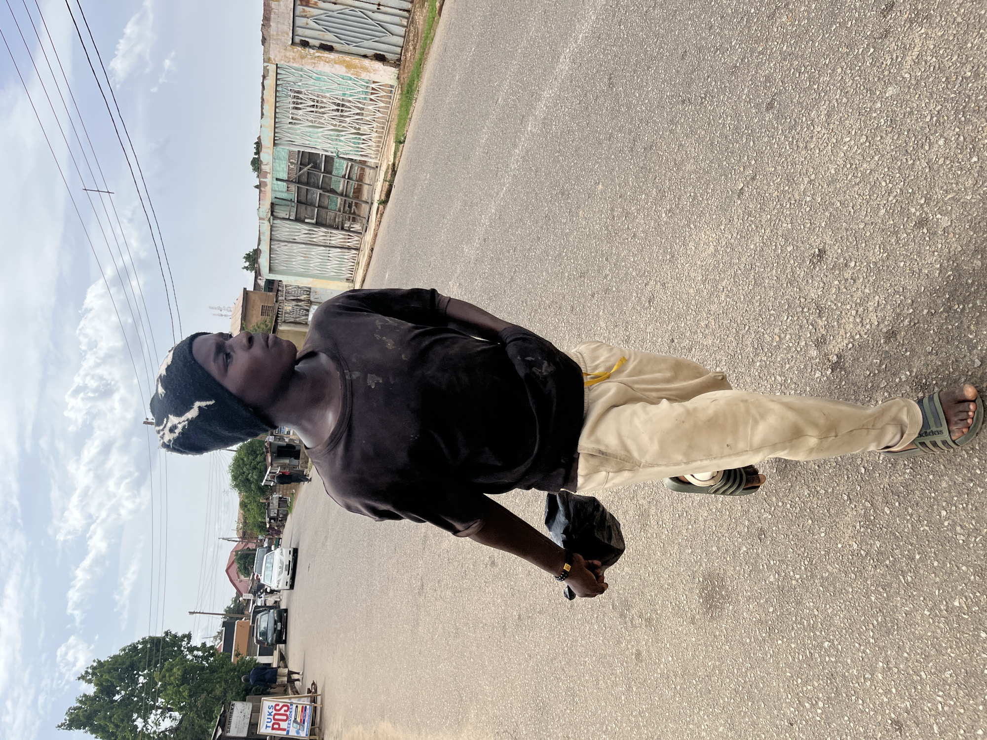 A man stands on a paved road in Jos, Nigeria, looking upwards. He wears a black, patterned beanie, a black t-shirt, and khaki trousers. He is also wearing black and grey Adidas sandals. He holds a black plastic bag in his right hand. In the background, there are buildings, vehicles, and trees. Overhead electrical wires stretch across the sky. The day appears overcast.