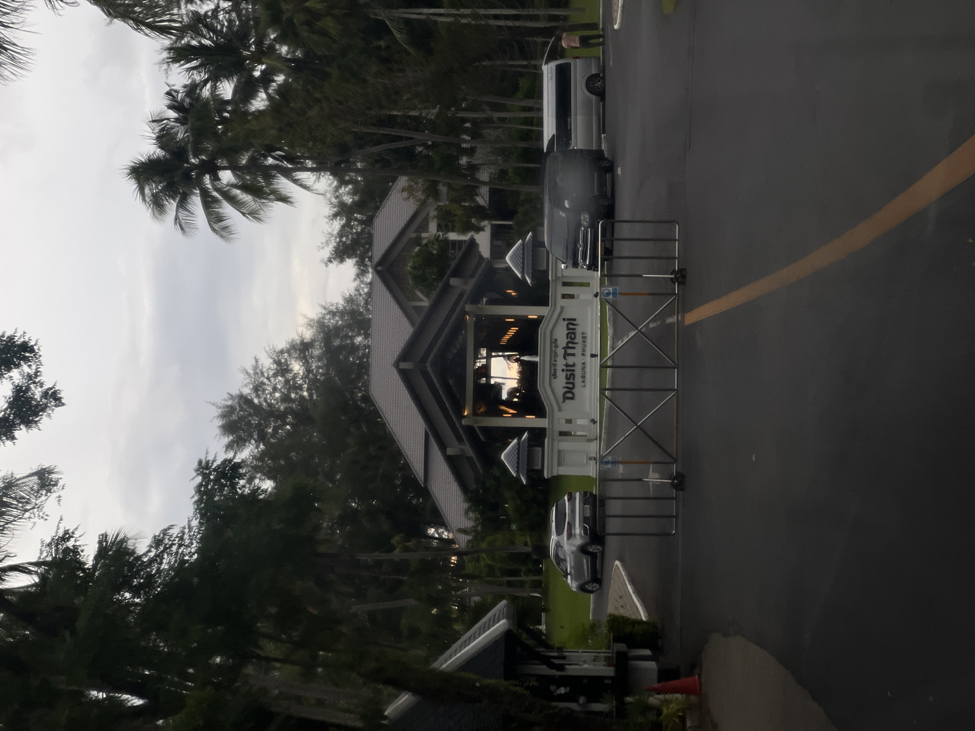 The image shows the entrance to a resort or hotel, likely in a tropical location, given the presence of palm trees. The main building has a distinctive, multi-tiered roof structure. A white gate with metal bars is positioned at the entrance, with a sign visible on a white structure behind it. The sign features text in both Thai and English, with "Dusit Thani" and "LAGUNA PHUKET" clearly legible.
Several vehicles are visible: a white van and a dark-colored van or SUV are parked on the asphalt road leading to the entrance. A silver sedan is also seen on the grounds. The scene appears to be set in the late afternoon or early evening, as indicated by the soft, diffused light and the overcast sky. The overall atmosphere is somewhat subdued, possibly due to the weather or the time of day. The environment is lush with greenery, with tall trees framing the scene and suggesting a warm climate. The perspective of the shot is from a slightly elevated angle, looking down towards the entrance.
