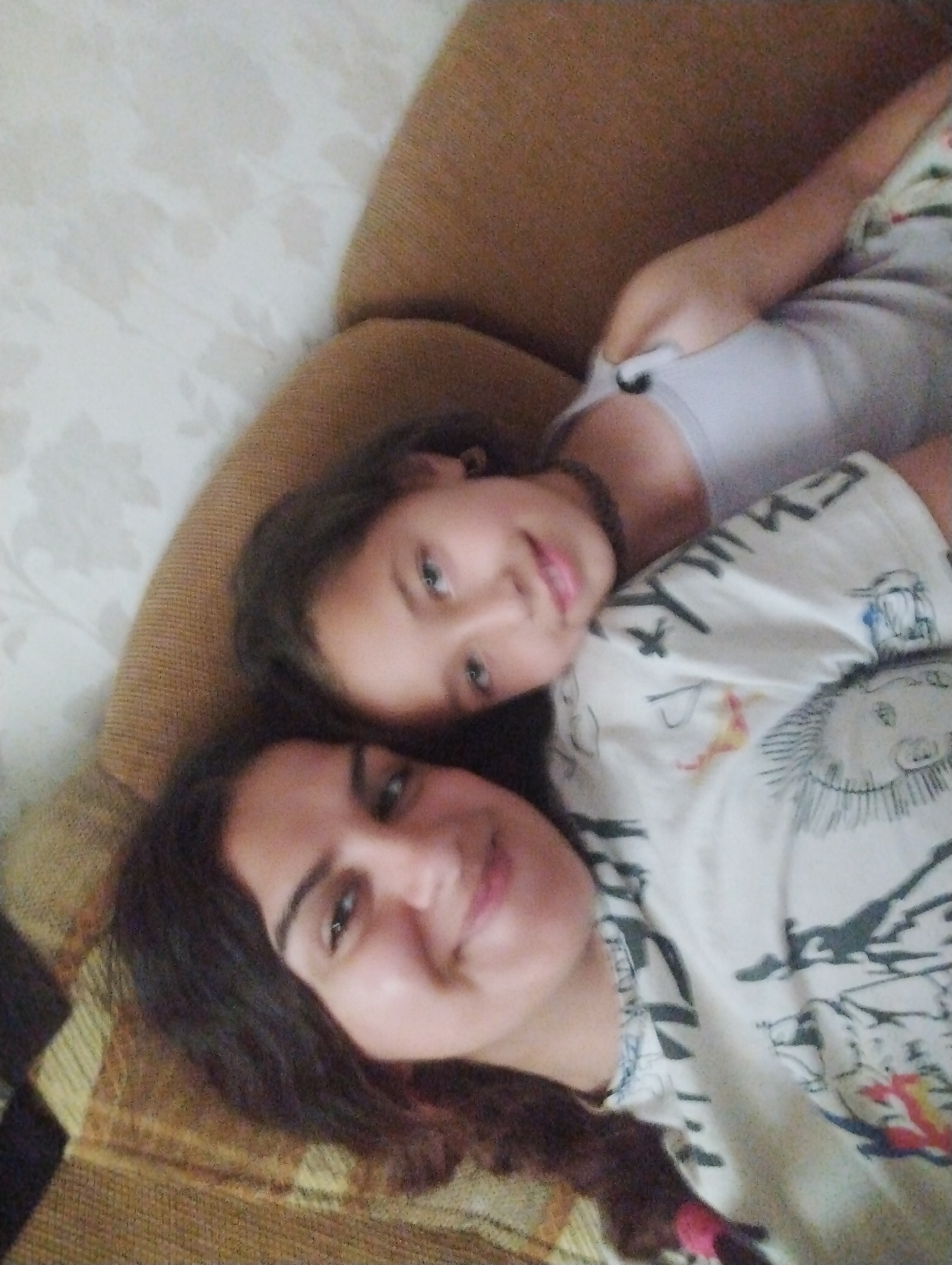Two individuals, appearing to be a mother and child, are lying down on a textured surface that could be a couch or bed. The child, positioned behind and slightly above the adult, is resting their head on the adult's shoulder. The adult is looking upwards and slightly to the side, with their mouth open. The child's face is partially visible, with their eyes looking towards the camera. They are both wearing casual clothing. The environment is dimly lit, suggesting an indoor setting. The image does not provide any specific information about the location in Hrishteni, Bulgaria.