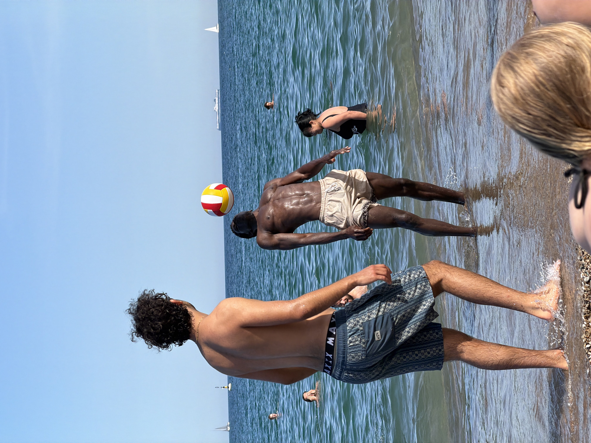 The image depicts a sunny day at a beach, likely in Barcelona, Spain, with people engaged in recreational activities in the Mediterranean Sea. In the foreground, a man with curly dark hair stands facing away from the camera, wearing patterned blue swim shorts. To his right, another man, who is darker-skinned, is in motion, with his body arched and head tilted back, appearing to be hitting a volleyball, which is visible in the air above him. This volleyball is striped with red, yellow, and white.
In the background, several other individuals are visible in the water. A woman in a black swimsuit is to the right of the man playing volleyball, and another woman with long dark hair is further out in the sea. Two men are also visible in the water to the left, with one appearing to shield his eyes.
The sea is a clear blue, with gentle waves reaching the shore. The beach itself is sandy with some pebbles. In the distance, several sailboats are scattered across the horizon, along with a large white ship. The sky is a clear, bright blue, indicating fair weather. The scene conveys a relaxed and active atmosphere of a summer day at the beach.