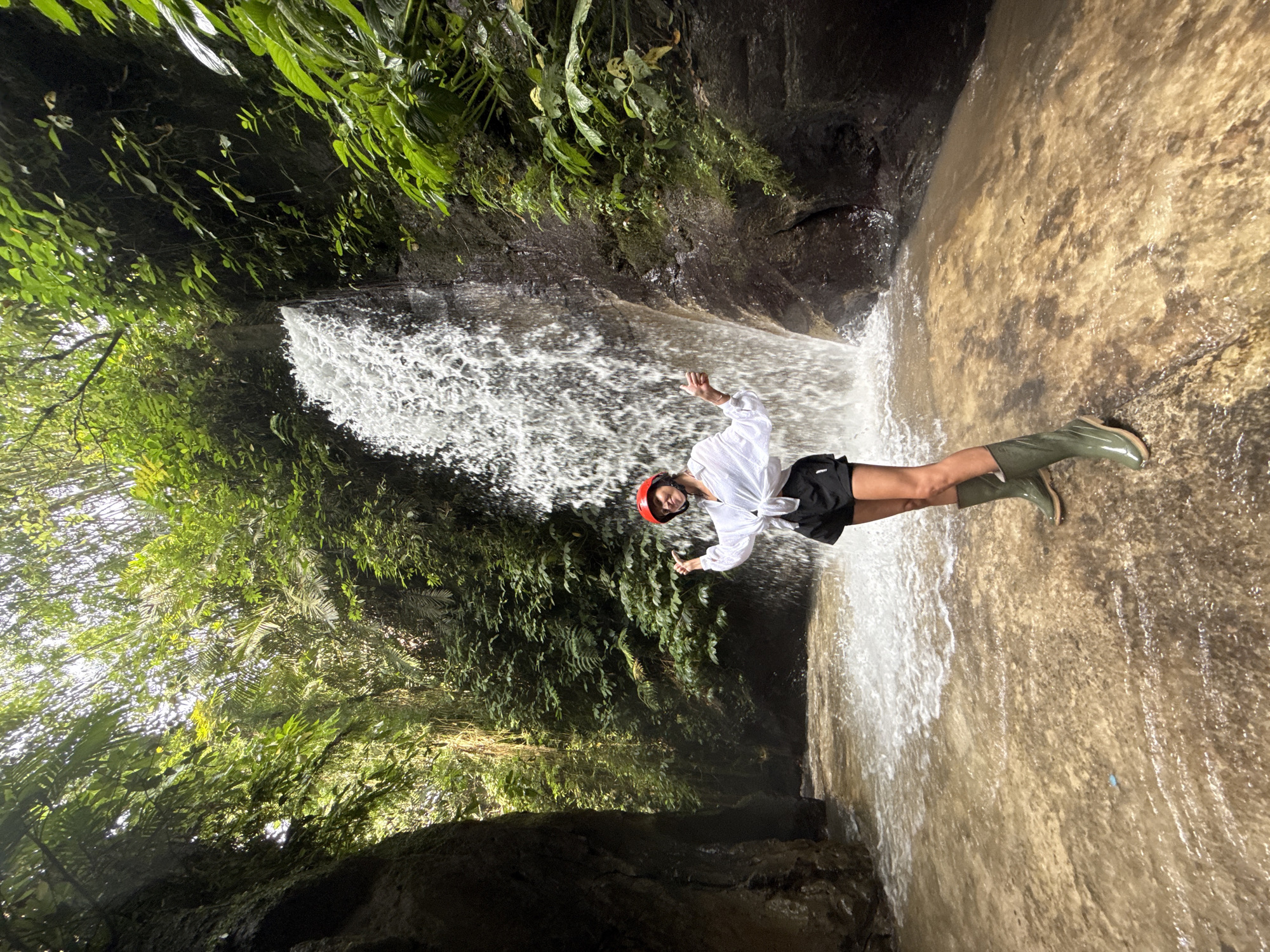 The image displays a young adult female standing in a shallow, brownish stream at the base of a waterfall in Payangan, Indonesia. The waterfall cascades down a dark, textured rock face on the right side of the frame. Dense, lush green tropical foliage, including trees and ferns, borders the waterfall and fills the upper and background areas, with sunlight filtering through the canopy. A large, dark rock wall forms a canyon-like structure on the left. The female wears a white long-sleeved shirt tied at the waist, black shorts, and tall olive-green rubber boots, complemented by a red helmet. She faces the camera with outstretched arms, palms upward, and a wide smile, positioned slightly to the left of the main water flow.