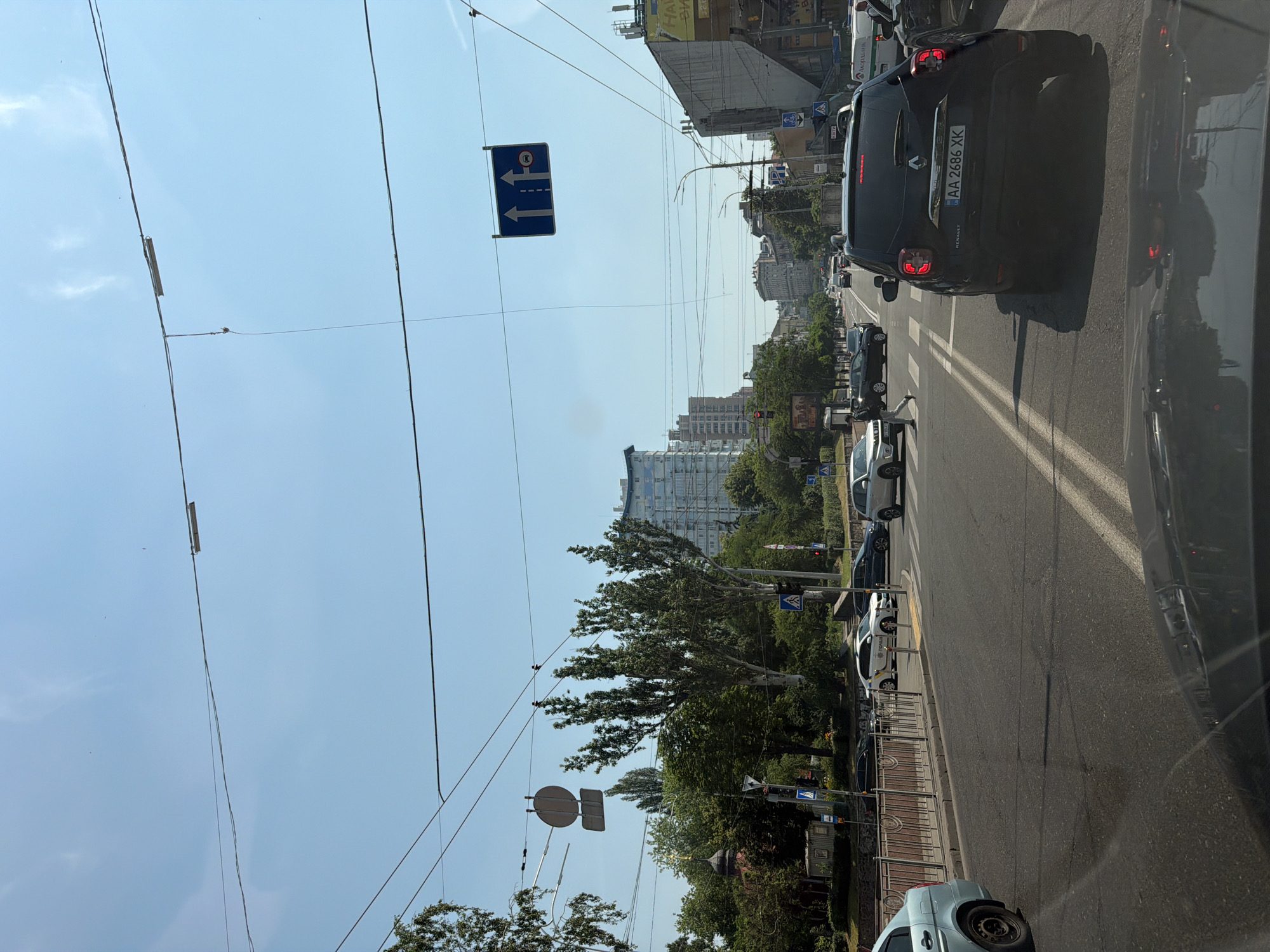 The image depicts a street scene in Kyiv, Ukraine, under a clear blue sky. The perspective is from inside a vehicle, looking forward. The street is paved and has multiple lanes, with traffic visible in all lanes. Several cars are present, including a black Renault Duster in the foreground with license plate "AA 2686 XK", a light blue car on the left, and various other vehicles further down the road. A pedestrian is crossing the street at a marked crosswalk.
On the left side of the street, there is a sidewalk with a metal railing, trees, and a small red building with a golden dome. A traffic light is visible on a pole, currently showing red. Further down the street, tall buildings with modern architecture are present. Overhead, power lines and a traffic sign displaying two upward arrows and a red circle with a diagonal line through it are strung across the street. On the right side of the street, there are more buildings, some appearing to be shops or commercial establishments, with signage.
The overall environment suggests an urban setting with active city life and traffic. The lighting indicates it is daytime, likely late morning or early afternoon, given the clear sky and shadows.