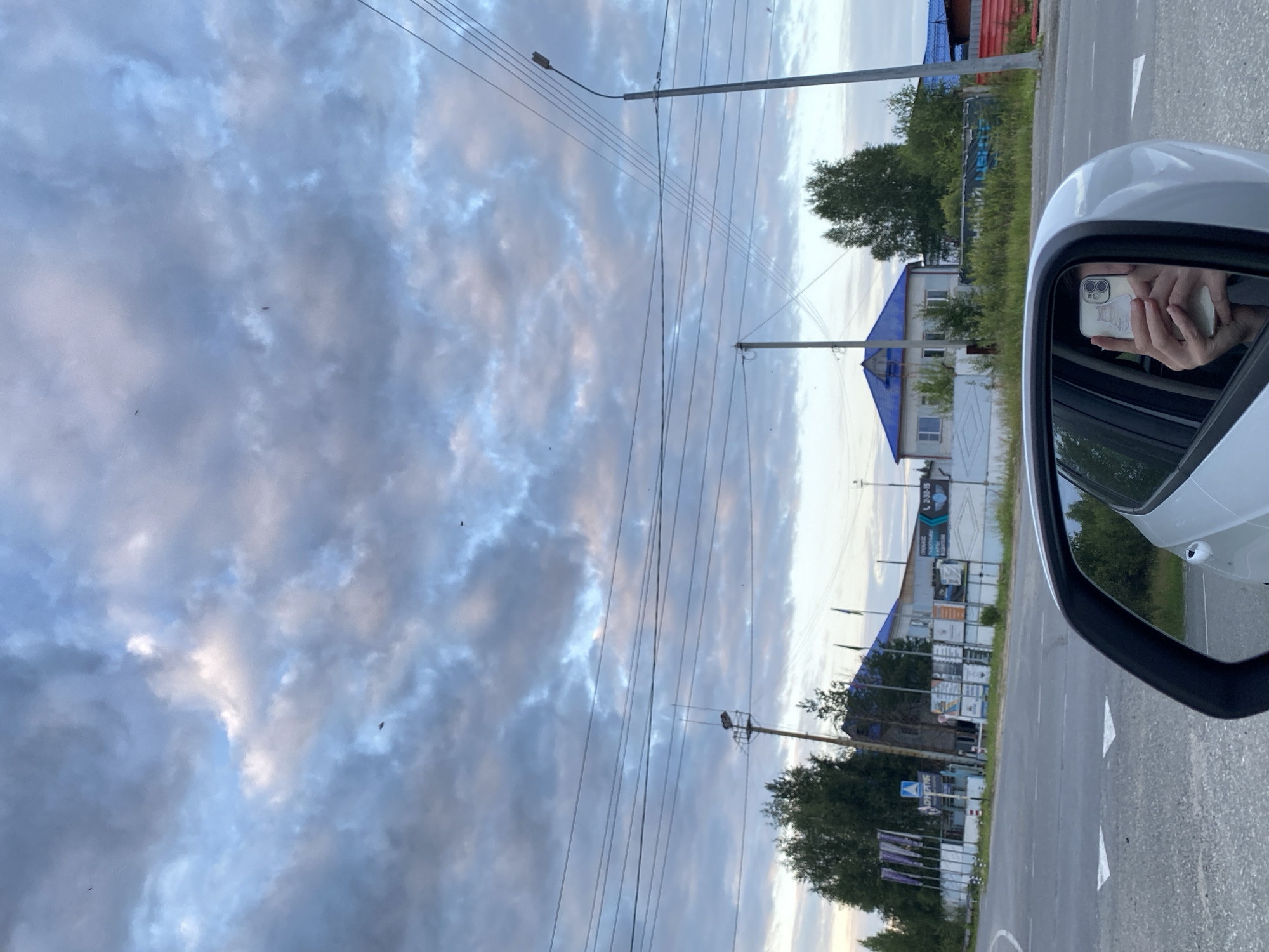 A person is taking a selfie in the side mirror of a white car. The person's hands are holding a light-colored smartphone, and their face is reflected in the phone's screen, which has a cat-themed case. The background is a street scene in Nyagan, Russia, with a road, buildings with blue roofs, trees, and utility poles. The sky is cloudy and overcast. There are also some signs and fences visible.