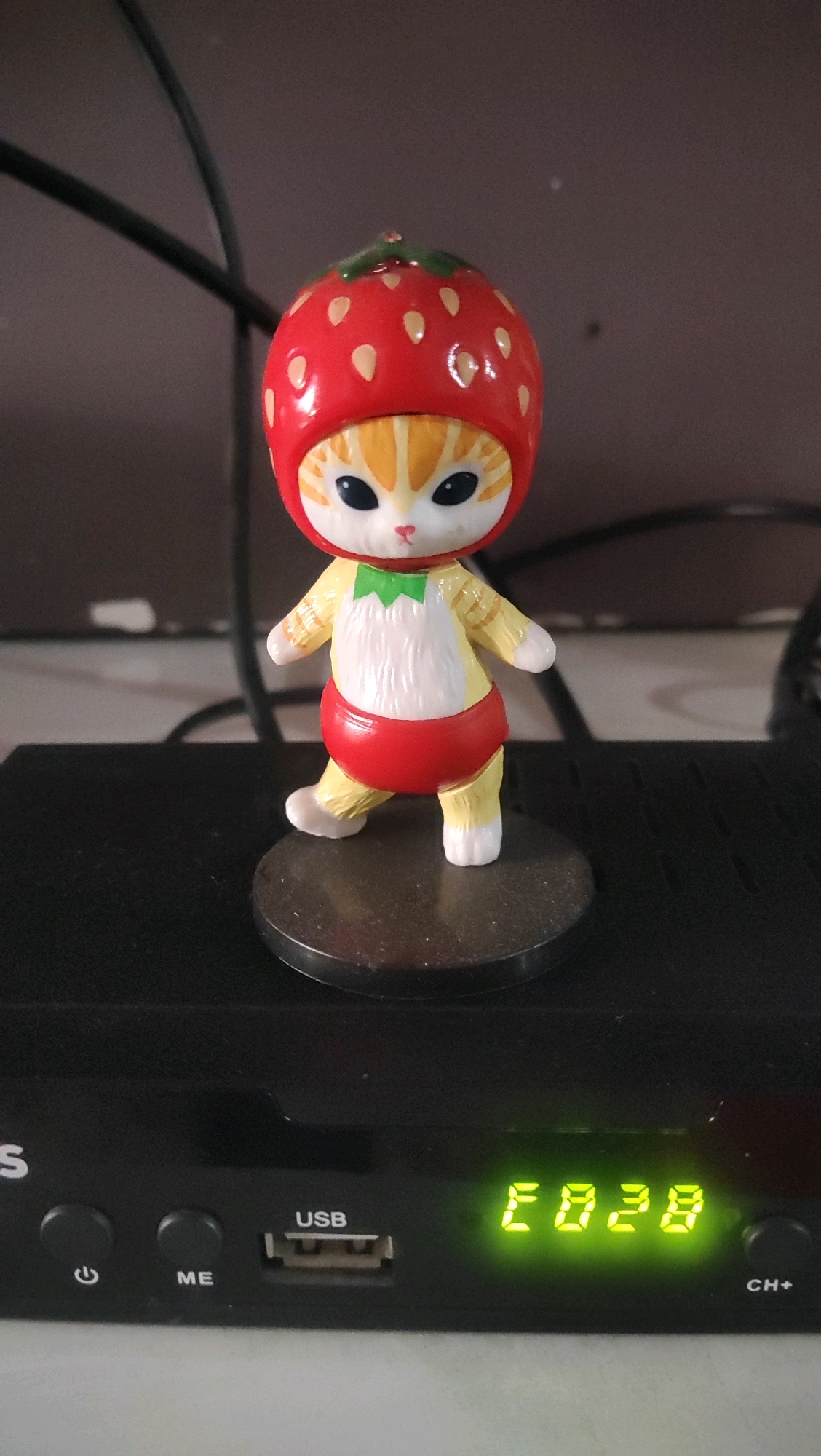 A close-up, eye-level, vertical shot shows a small, novelty figurine of a cartoon cat wearing a strawberry-shaped hat. The figurine is positioned centrally on top of a black electronic device, likely a satellite receiver, which is sitting on a light-colored surface. The cat figurine is predominantly yellow and white, with orange tabby markings on its face and body. It has large black eyes and a small pink nose. It is wearing a green bow tie and a red diaper-like garment. The figurine is mounted on a small black circular base. The electronic device below has several buttons and a USB port. To the right of the USB port, a green digital display shows the characters "E028". The background is dark and out of focus, with some dark cables visible. This image was taken in the city of Karangwangkal, Indonesia.