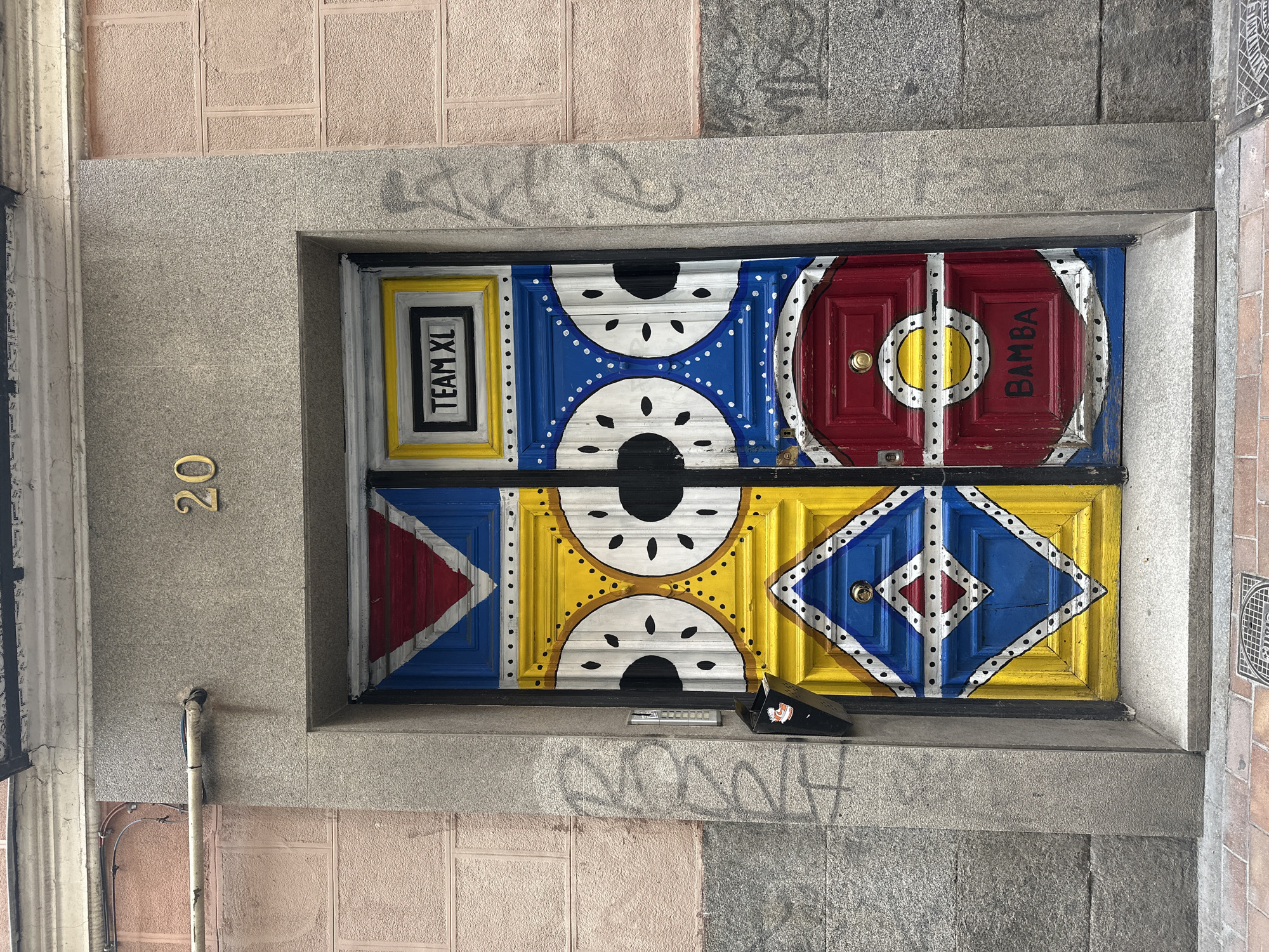 The image shows a vibrant, heavily decorated double door in Madrid, Spain, located at number 20. The door is divided into four panels, each with a distinct colorful design. The top left panel features a red triangle pointing down on a blue background with white borders. The top right panel has a rectangular black and white sign that reads "TEAM XL". The middle left panel is yellow with a white and black circular design, resembling two eyes with black pupils and white dots as eyelashes. The middle right panel is blue and has a similar circular design as the middle left panel. The bottom left panel is yellow and features a large blue diamond shape with a red diamond inside and white borders with black dots. The bottom right panel is red and has a circular design with a yellow circle in the center, and below that, the word "BAMBA" is written in black. The door is set within a gray stone facade. To the left of the door, a black mailbox is attached to the wall. The surrounding building walls are textured, one appearing pinkish and the other a speckled gray. Graffiti is visible on the gray facade and the pink wall. The ground in front of the door is paved with bricks and a manhole cover is present.