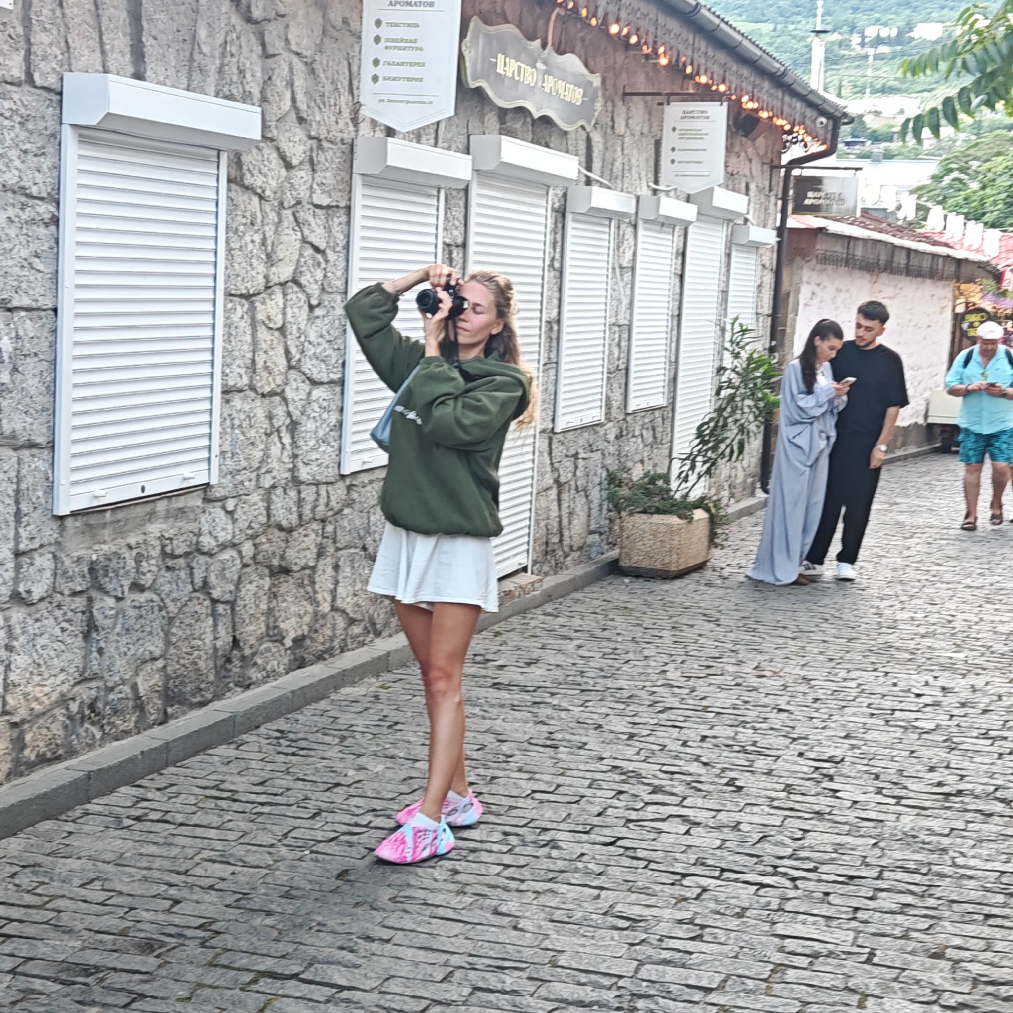 A young woman with long, wavy blonde hair stands in the middle of a cobblestone street, holding a black camera to her face and taking a picture. She is wearing a loose, olive-green hooded sweatshirt and a short, white skirt. Her feet are adorned with bright pink and blue athletic shoes. Behind her, a stone building with multiple windows covered by white roller shutters runs along the street. Above the windows, ornate signs with Cyrillic lettering are visible, one of which reads "ЦАРСТВО АРОМАТОВ" (Kingdom of Aromas). To the right of the woman, a couple is standing; the man, wearing a black t-shirt and pants, looks at his phone, while the woman next to him, dressed in a long, light blue flowing garment, is also engrossed in her phone. Further down the street on the right, a man wearing a light blue short-sleeved shirt and patterned swim trunks is walking away from the camera, also looking at his phone. The street is lined with potted plants, and the background shows a hillside with buildings and greenery, suggesting a town or village setting. The overall scene appears to be in an outdoor commercial or pedestrian area.