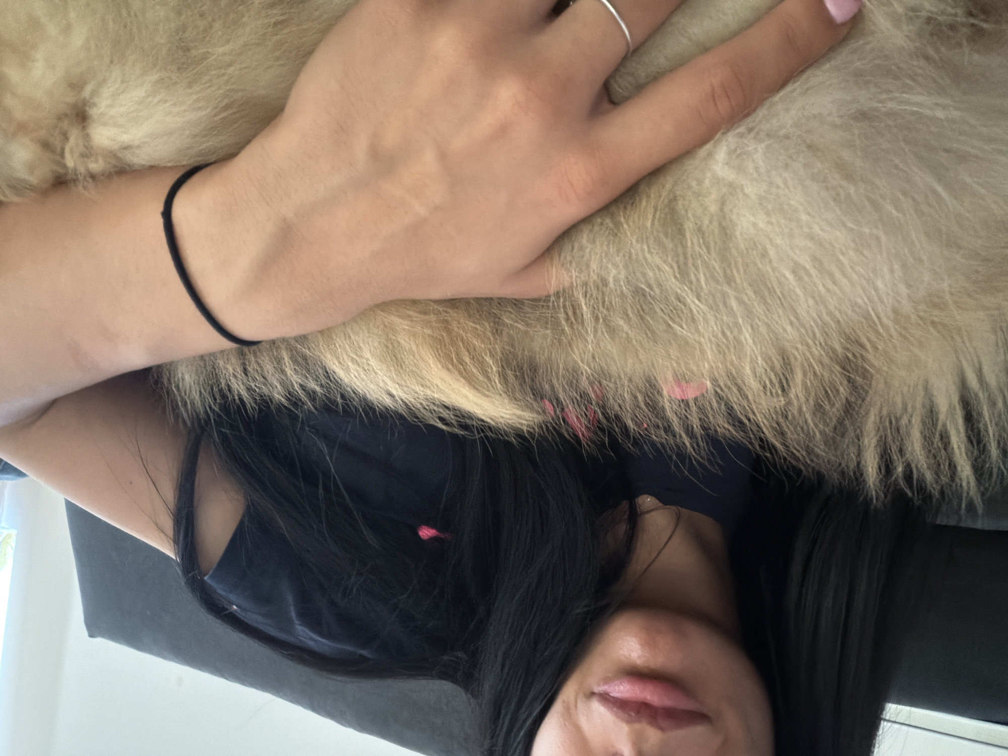 The image depicts a woman lying on a dark surface, possibly a couch, with her dark hair spread around her face. She is wearing a black top. Her hand rests on a light brown, furry object, potentially a dog or a fur blanket. She has a silver ring on one finger and a black hair tie around her wrist. Her lips are visible in the lower part of the image. There is nothing in the image to identify it as being in Shumen, Bulgaria.