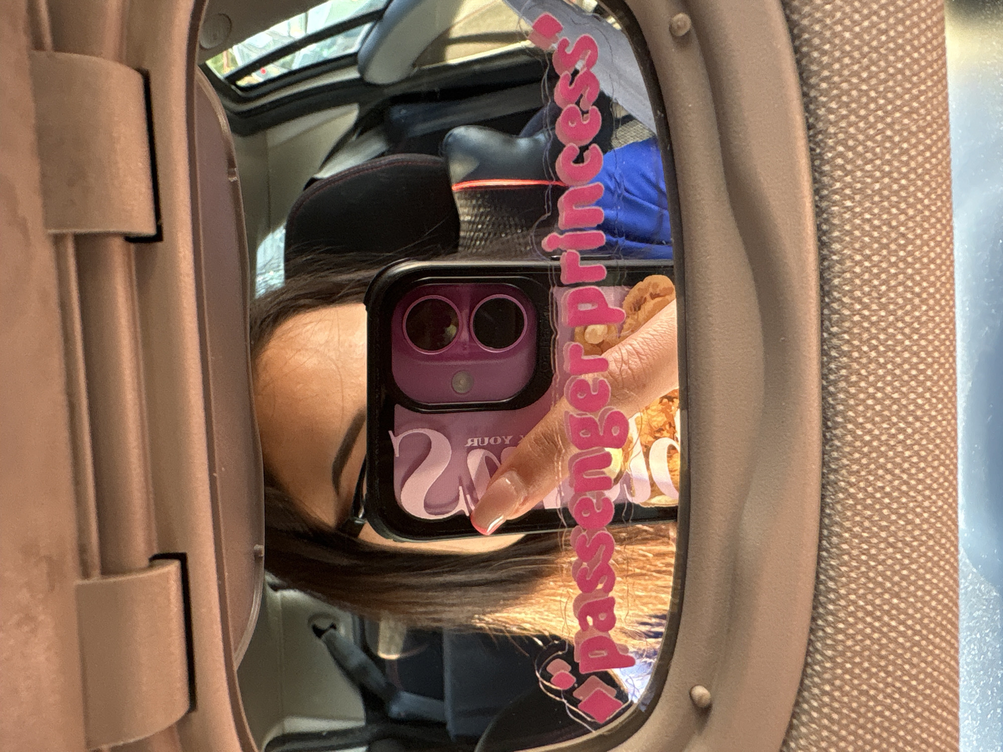 A person is taking a mirror selfie in a car. The person's face is partially visible, and they are holding up a phone with a pink case. The mirror has a pink sticker on it that says "passenger princess". The interior of the car is visible, including the seats and the roof lining. The car appears to be parked. The scene is an interior shot of a car, no specific location in Vratsa, Bulgaria is identifiable.