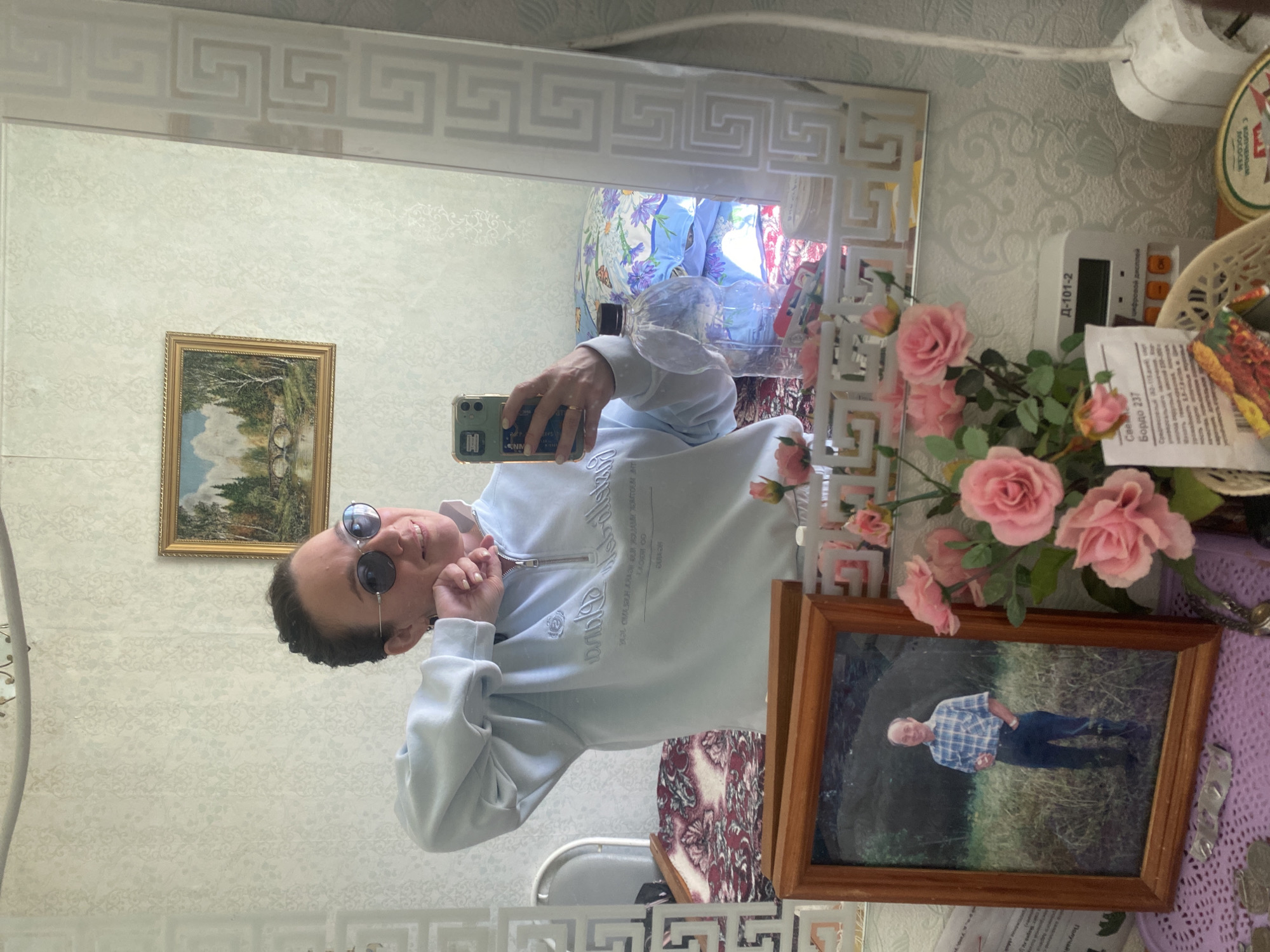 A mirror selfie taken in what appears to be a home interior.
**Person:** A person wearing round sunglasses and a light-colored zipped sweatshirt is taking the selfie. Their face is visible, and they are holding up a smartphone.
**Subjects/Objects:**
* Mirror: A large mirror with a decorative patterned frame.
* Wall: The wall behind the mirror is covered with patterned wallpaper.
* Decorations: A framed picture with a portrait, artificial pink roses, and a basket are visible on a surface beside the mirror.
* Other: There are also items such as a device (possibly an electronic control panel), and some paperwork.
**Actions:**
* The person is holding up their phone to take a picture of themselves in the mirror.
**Environment:**
* The setting appears to be inside a home with a traditionally decorated interior.
**Location:** This is not knowable based on the image. Узяр, Russia is mentioned in the instructions only.