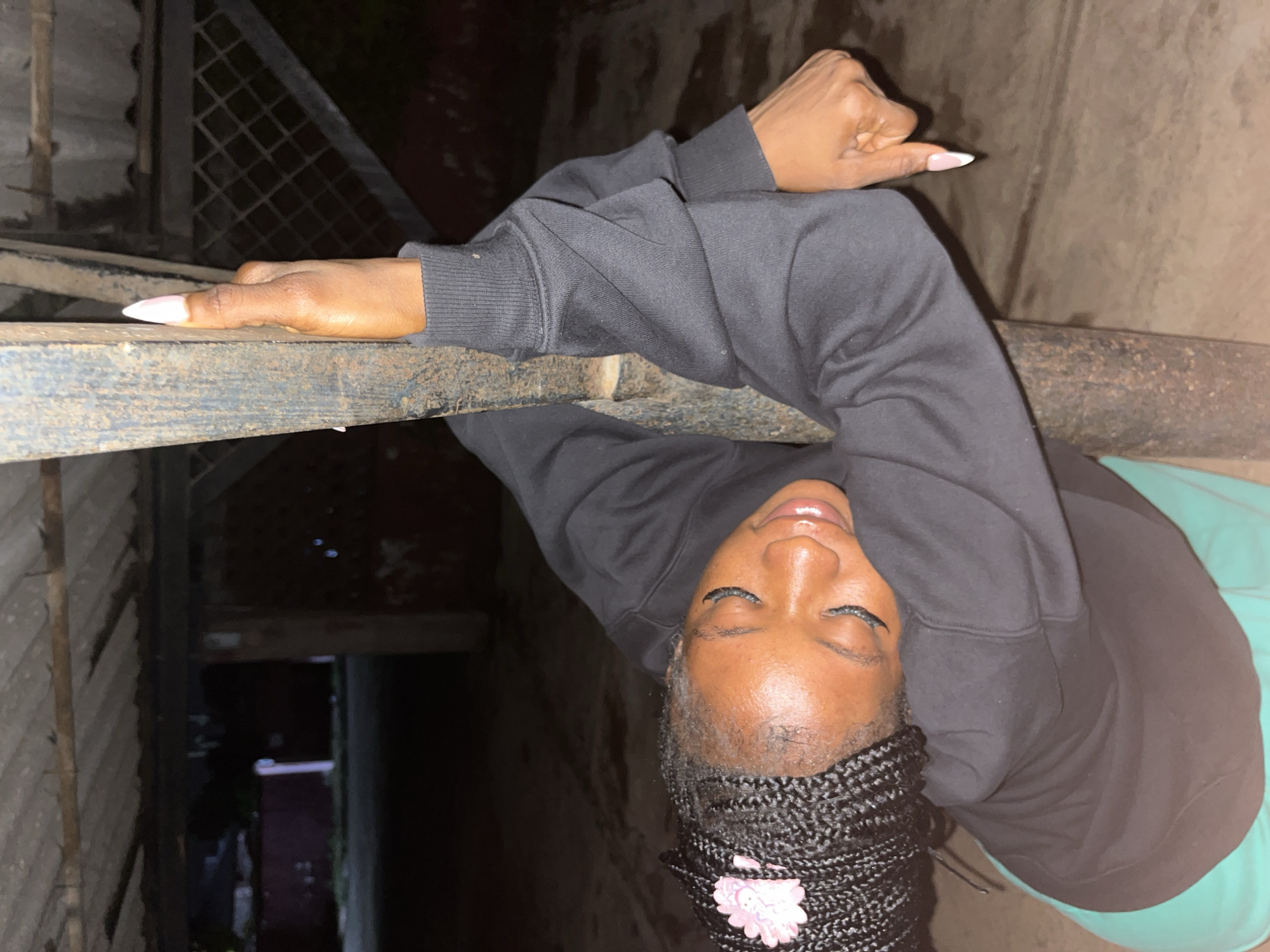 A young Black woman with braided hair, adorned with a pink flower, rests her head and arms on a rough, vertical metal pole. She is wearing a black sweatshirt and a teal garment, possibly pants or a skirt. Her eyes are closed, and she appears to be in a relaxed or sleepy state. Her fingernails are long and painted white with pink tips. The background suggests an outdoor, possibly covered area, with a corrugated roof overhead and a dimly lit, gritty ground surface. The overall impression is one of a candid, personal moment captured in an urban environment. The location is a public or semi-public space in Jos, Nigeria, likely an outdoor shelter or covered parking area, given the visible corrugated roof and the rough, uneven ground.