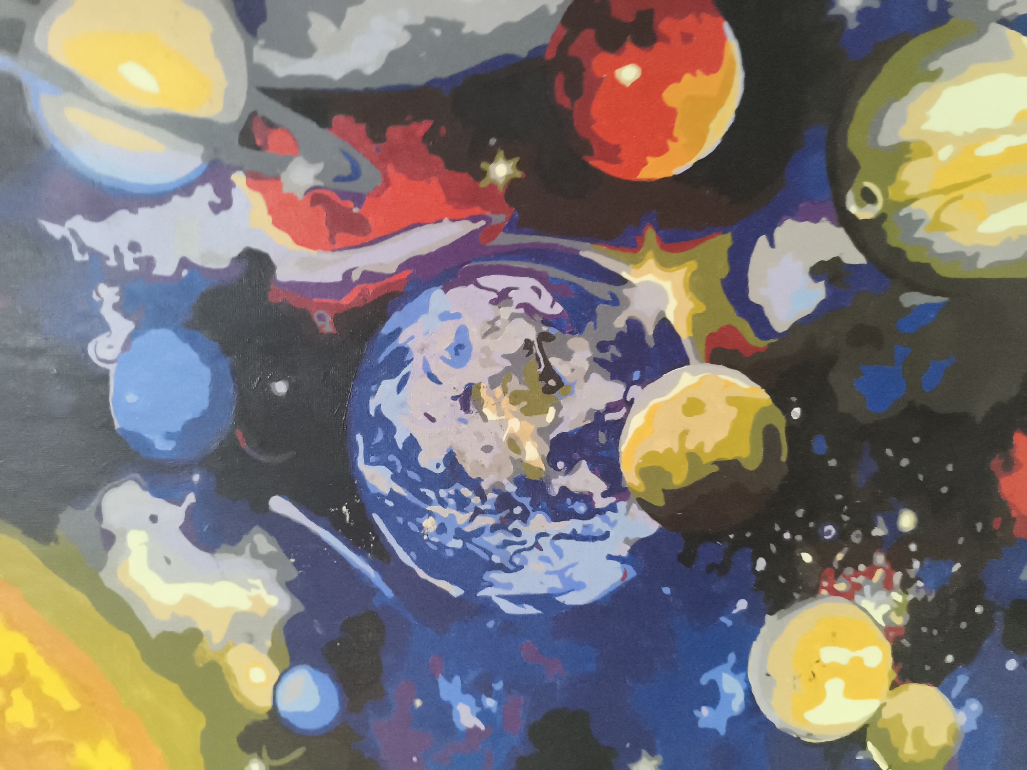 The image is a painting depicting a stylized version of outer space with various planets. Prominent planets include a blue and white planet resembling Earth, a red planet possibly representing Mars, and several yellow and green planets. There are also star-like shapes and nebulous clouds painted in the background. The style is amateurish and likely a paint-by-numbers project. The scene has no connection to the city of Nowy Tomyśl, Poland.