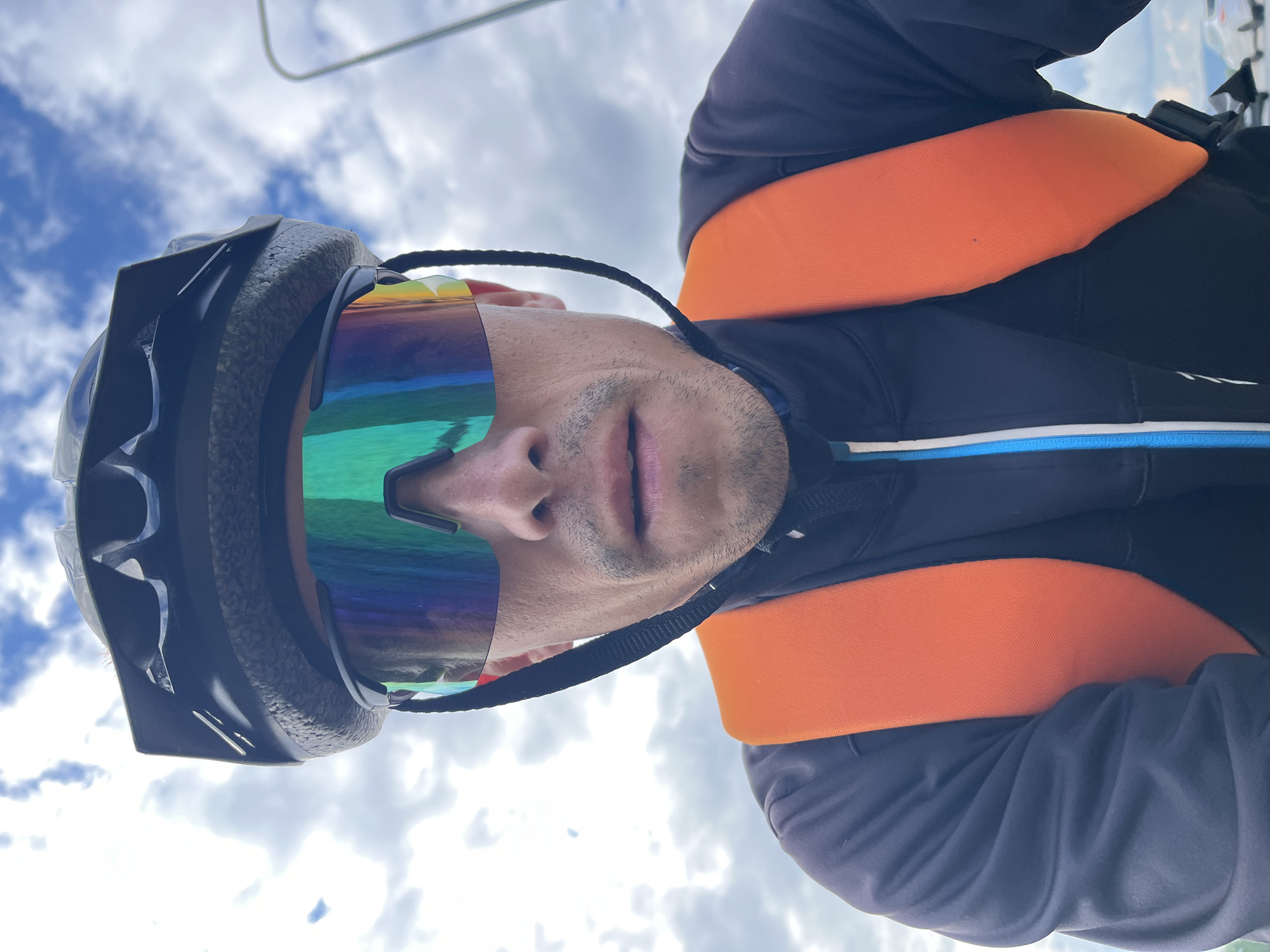A close-up, upward-angled portrait captures a person wearing outdoor sports gear against a bright, cloudy sky. The individual, appearing male, is shown from the chin upwards, wearing a black helmet with a textured inner lining and large, highly reflective sports sunglasses that predominantly cover their eyes and forehead. These iridescent sunglasses exhibit a striking gradient of green, blue, purple, and orange hues. Below the sunglasses, the person's nose, mouth, and chin, featuring light stubble, are visible, displaying a neutral expression.
The person is wearing a black jacket, and a prominent, wide orange band or strap wraps horizontally across their torso, suggesting it might be part of a life vest, safety harness, or specialized sports equipment. A thin blue and white stripe is also visible on the lower portion of the black jacket. The background is a bright, partly cloudy sky, indicating an outdoor, daytime setting. No visible text is present in the image. The attire suggests the person is engaged in an active outdoor sport or recreational activity, such as cycling, skiing, or water sports, aligning with the geographical context of Chaikovsky, Russia.