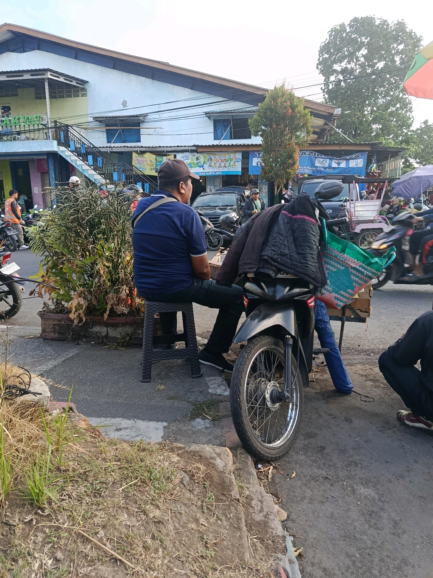 A man wearing a blue t-shirt and a dark baseball cap sits on a black plastic stool facing away from the camera, his left shoulder towards it. He is positioned next to a black motorcycle. The motorcycle has several items draped over its rear, including a dark jacket and a woven green and red bag. A blue cloth or tarp hangs from a stand behind the motorcycle, with a portion of it draped over the motorcycle seat.
The motorcycle is parked on a paved street, which appears wet in places, suggesting recent rain. To the left of the motorcycle is a large, overgrown potted plant. In the background, a multi-story building with a light blue facade and a light green section is visible. Signs with text are present on the building and adjacent structures.
Several other motorcycles and a few cars are visible further down the street, indicating a bustling urban or semi-urban environment. People can be seen in the background, some near motorcycles and others by the buildings. The overall scene suggests a street-side marketplace or a busy road in Bululawang, Indonesia. The sky is overcast, and the lighting suggests it is daytime.