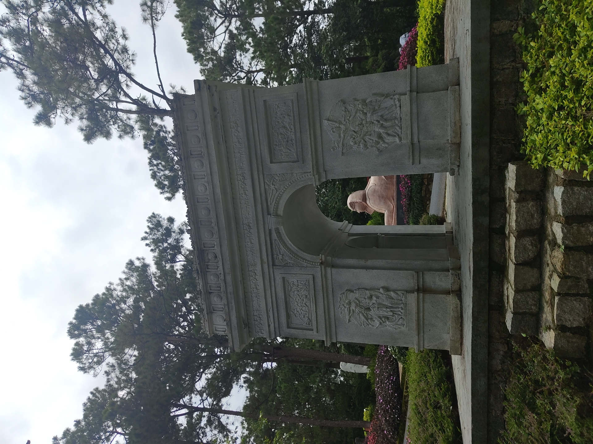 The image captures a stone archway in a garden setting, likely in Lâm Viên - Đà Lạt, Vietnam. The archway is adorned with bas-relief carvings on its pillars, depicting figures and scenes. Through the arch, a large statue of a sphinx-like creature is visible, its body facing left and its head looking forward. The creature is a light brown color and appears to be made of stone or a similar material. Surrounding the archway are lush green bushes and flowerbeds with pink and purple blossoms. Tall pine trees are scattered throughout the background, and the sky is overcast. Stone steps lead up to the platform in front of the archway, and the ground is paved with stone slabs. The overall atmosphere suggests a historical or memorial site within a park.