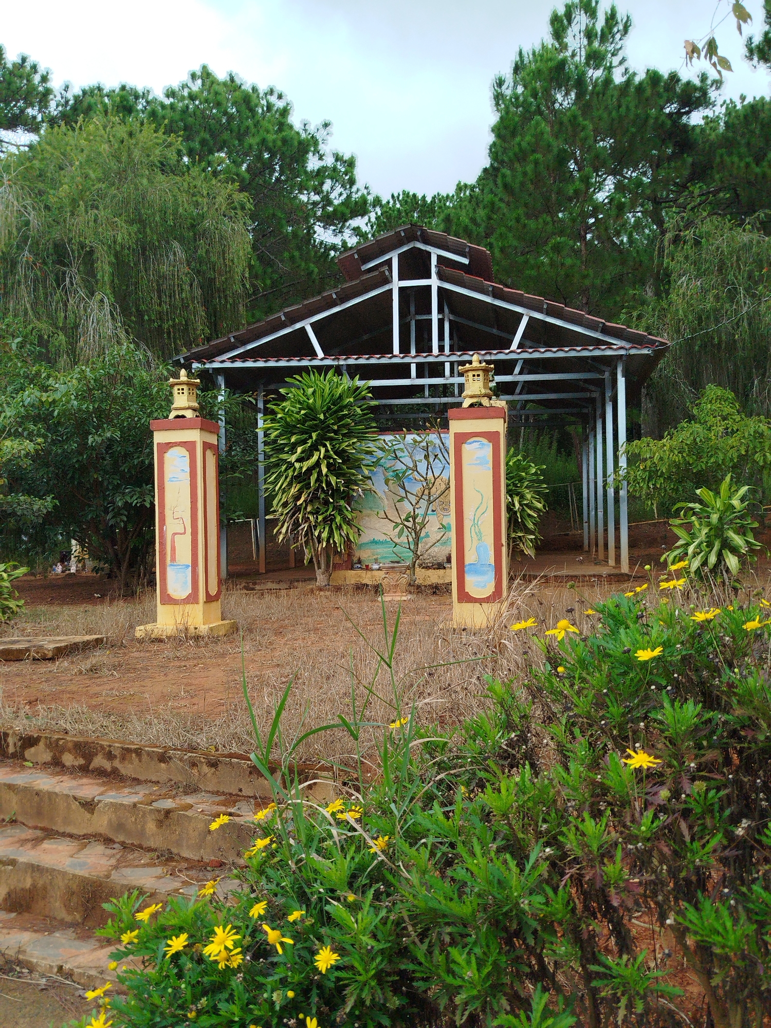A wide shot captures an outdoor pavilion in a park-like setting, likely in Lam Vien - Da Lat, Vietnam, given the vegetation and architectural style. The pavilion features a metal frame with a tiled roof, open on all sides, and appears to be set on red dirt. Two decorative pillars flank the entrance, painted in yellow and red, with simple artwork depicting a tree and blue sky. Behind the pillars, a mural of a stylized tree with a blue base is visible on the pavilion's back wall. Lush green trees, including what appears to be a weeping willow, surround the pavilion. In the foreground, a cluster of bright yellow daisies blooms amongst dry grass and some weathered concrete steps leading up to the structure. The sky is overcast, suggesting a cloudy day. There are no people or animals visible in the image.