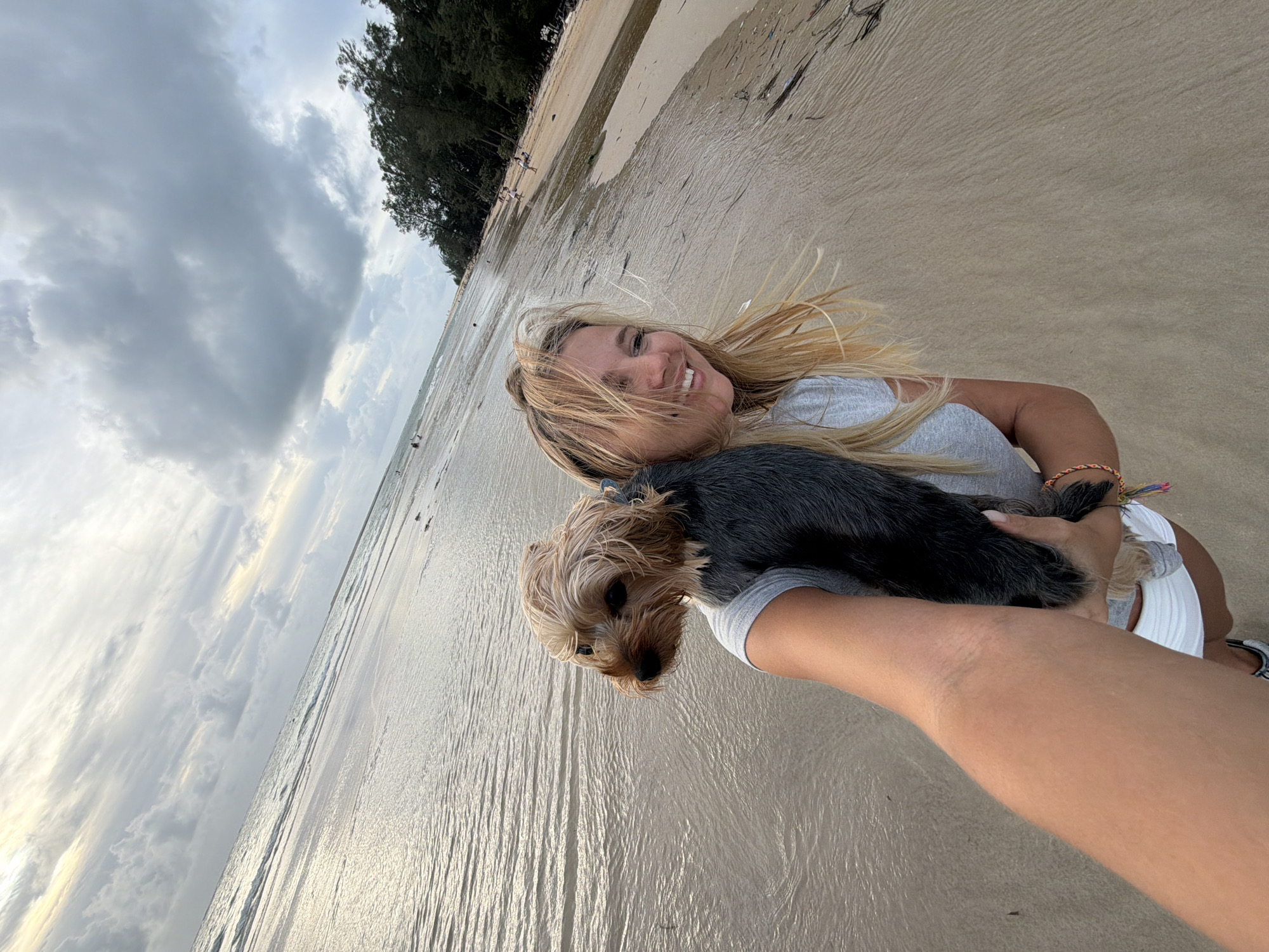 A selfie is taken on a beach in Sakhu, Thailand, with a woman holding a Yorkshire terrier. The woman has blonde hair and is wearing a gray t-shirt and white shorts. She is smiling at the camera while the dog, with its tan fur and dark eyes, is perched on her shoulder, looking to the left. The beach has wet sand with ripples from the receding tide. In the background, the sea stretches towards the horizon under a cloudy sky. Trees line the right side of the beach, and a few people are visible in the distance. The overall lighting suggests it is late afternoon or early evening.