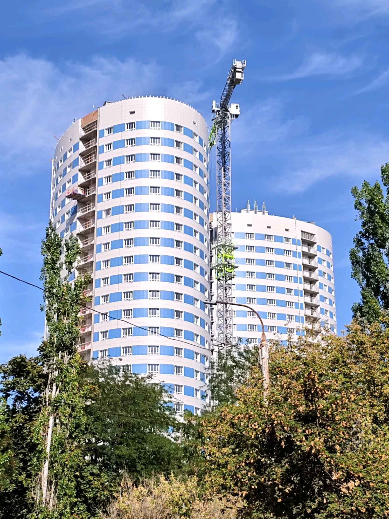 A construction crane is positioned next to a modern, curved, multi-story building with blue and white panels. The building appears to be under construction, with scaffolding visible on its side. Trees and foliage partially obscure the lower portion of the building. The sky above is blue with scattered white clouds. The scene is outdoors.