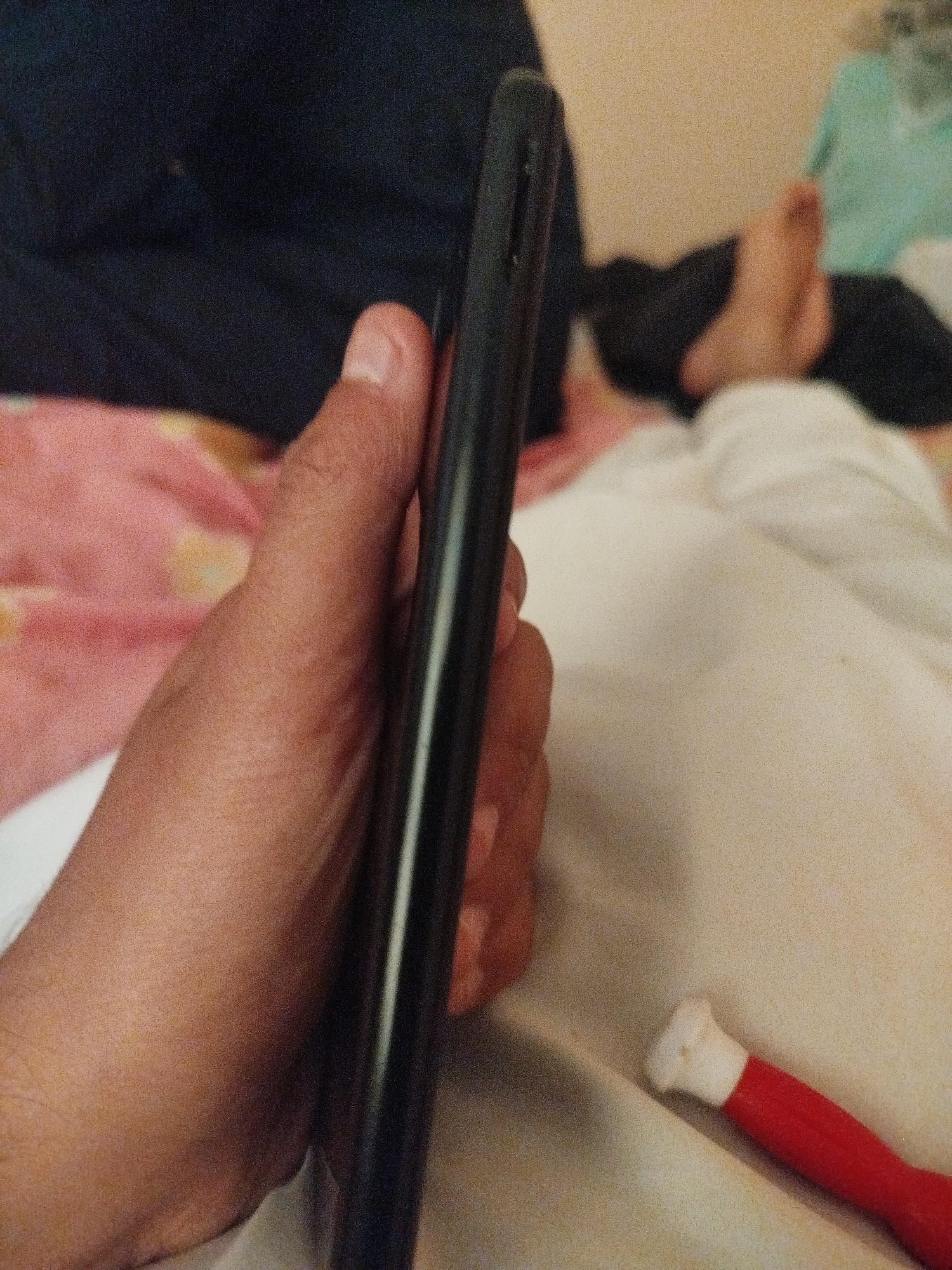 The image is a close-up shot of a person holding a dark-colored mobile phone. The phone is positioned vertically, and the person's hand is wrapped around its side. The background appears to be a domestic setting, with soft furnishings like blankets and a pillow visible. In the lower right corner of the image, a small, red and white object, possibly a toy or a piece of clothing, is partially visible. The lighting is dim, and the image has a grainy texture. The scene is indoors, and the overall impression is casual and informal. There is no discernible indication of the city of Lassan, Pakistan, within the image.