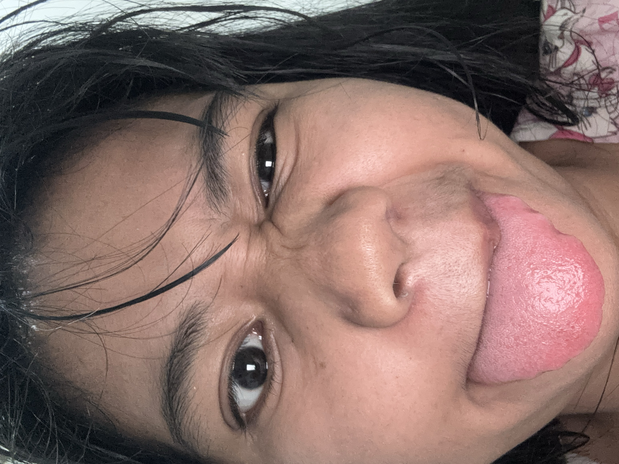 A close-up, forward-facing, medium-skinned individual is captured in a selfie. The person is making a face, with their tongue sticking out and their eyes squinted. Their dark hair is wet and falling across their forehead. The background is plain and light-colored, suggesting an indoor setting in North Jakarta, Indonesia.