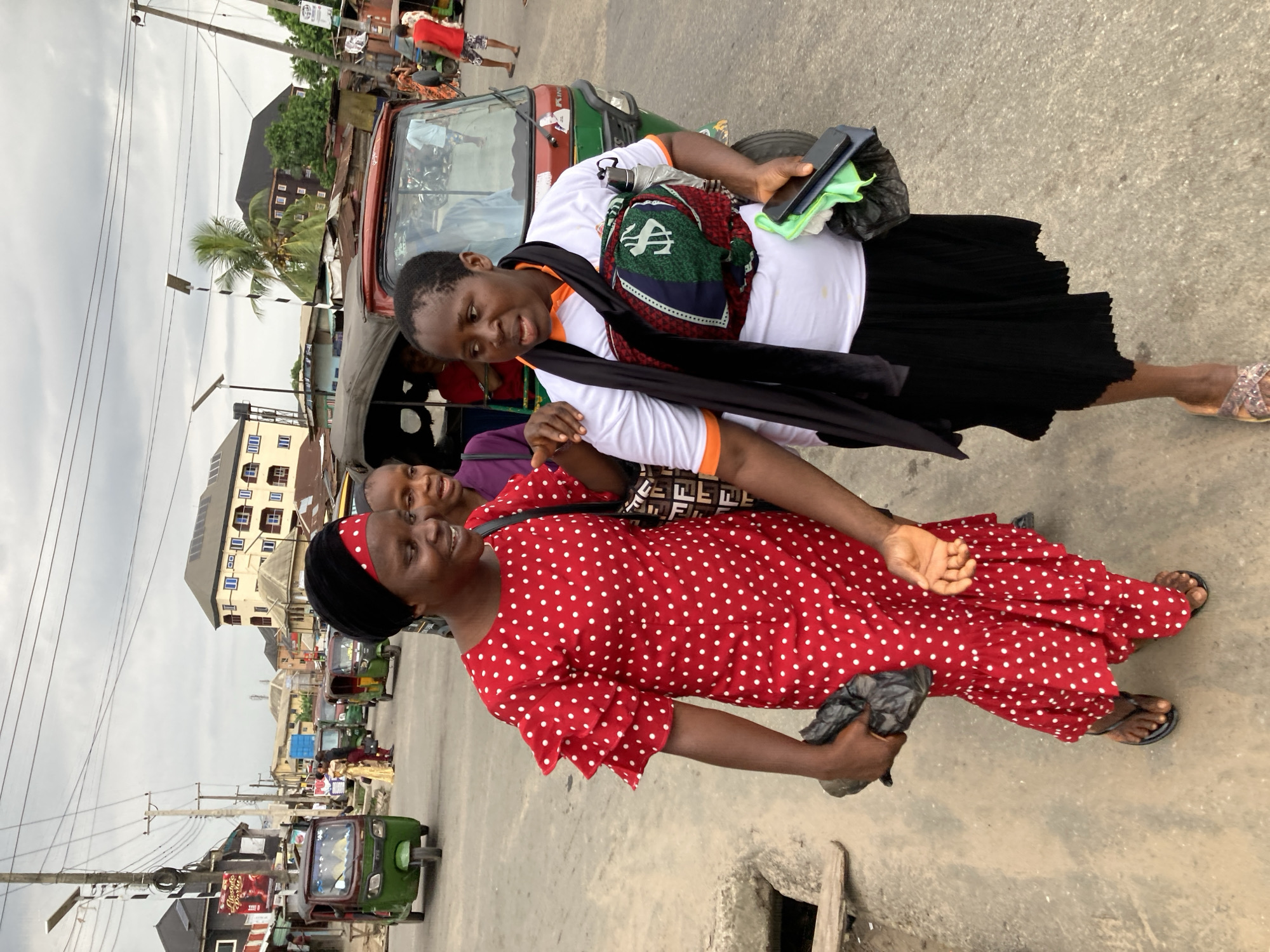 Two women stand close together on a paved street in what appears to be Aba, Nigeria. One woman, in the foreground, wears a red dress with white polka dots and a black head covering. She is carrying a black plastic bag. Behind her, another woman is dressed in a white shirt with orange trim, a black skirt, and carries a patterned shoulder bag. She is holding a phone and a green cloth. A three-wheeled vehicle, commonly known as a keke, is visible behind them. The street scene includes other kekes, buildings, utility poles, and a palm tree in the background. The sky is overcast.