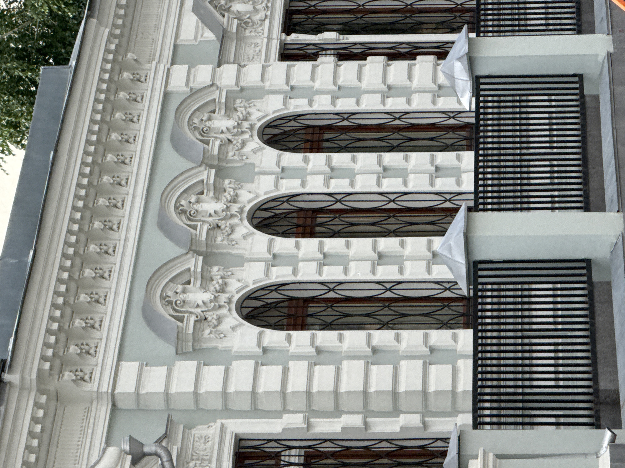 This image shows the facade of a building with classical architectural elements. The building is painted a light blue-grey with white decorative moldings and pilasters. Above the windows, there is a frieze with repeating decorative motifs and a cornice with dentil molding. The windows themselves are arched and feature dark metal grilles. In the foreground, there is a black metal fence and light-colored pillars topped with decorative caps. The scene appears to be located in Moscow, Russia, as evidenced by the architectural style which is common in the city's historic districts. There are no people visible in the image. The overall impression is one of a well-maintained historic building.
