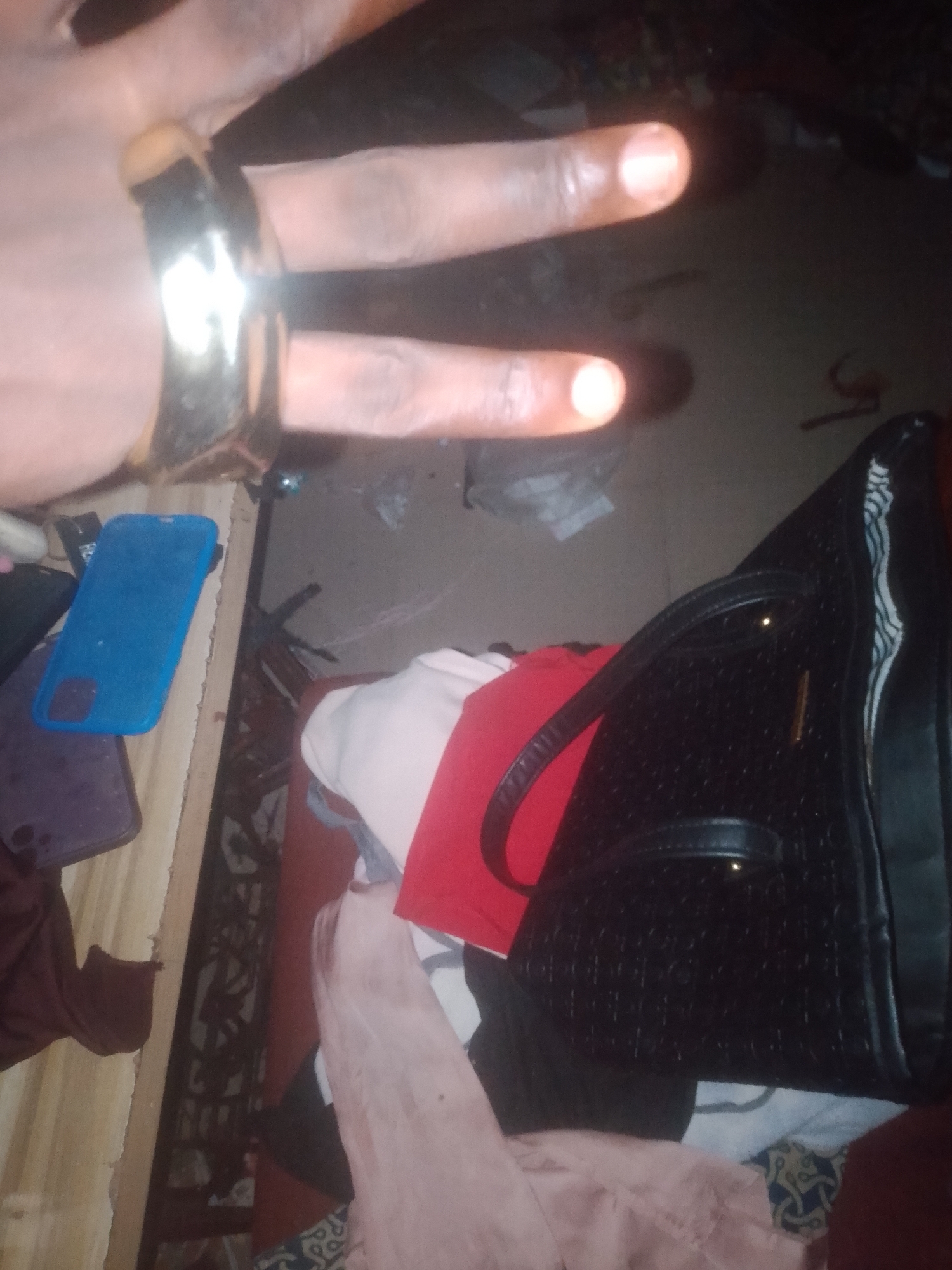 A person's hand, adorned with a gold ring, is held up with two fingers extended in a peace sign. In the background, a black textured handbag rests on a surface cluttered with various clothing items, including a bright red shirt and a blue phone case. The scene appears to be an indoor setting, possibly a room, with a dimly lit environment. The image does not provide any specific details to indicate a location within the city of Jauro Yinu, Nigeria.