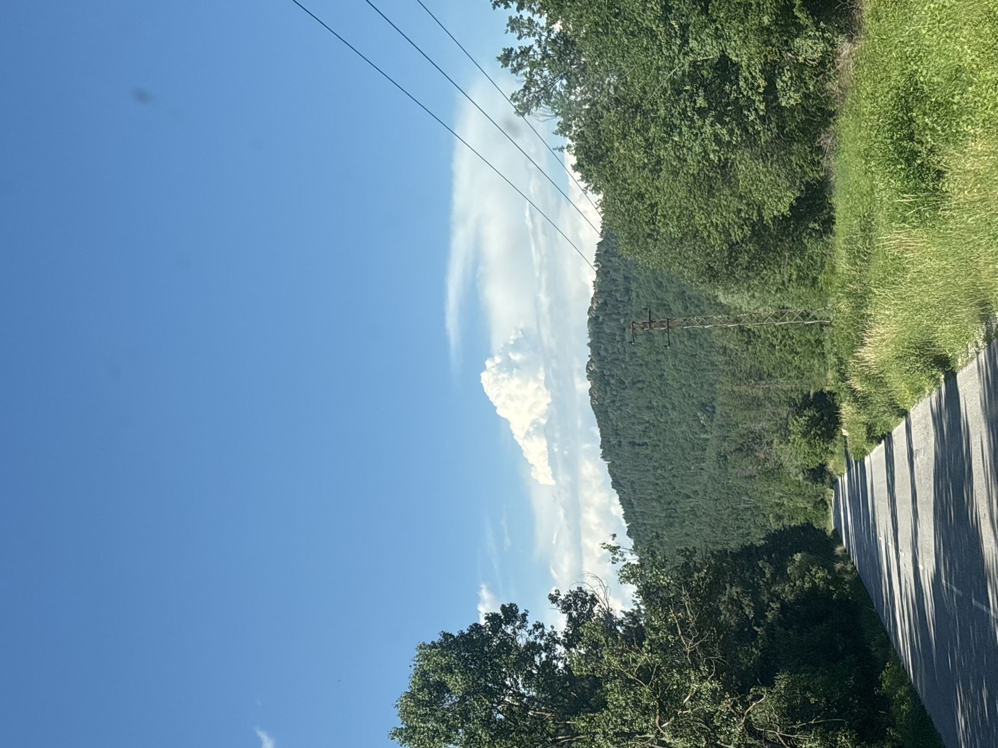 The image depicts a scenic view of a paved road winding through a lush, green landscape. The road is bordered by dense trees and tall grass, with shadows cast across its surface, suggesting it is a sunny day. In the background, a large, tree-covered hill rises, its peak dotted with rocky outcrops. A distinct cumulus cloud formation floats above the hill against a clear blue sky. The presence of power lines strung between poles indicates that this is a road in or near a populated area, likely a rural one. This scene is consistent with the natural beauty and terrain found near Topolovgrad, Bulgaria, a region known for its rolling hills and verdant forests. No people or specific actions are visible in the image.