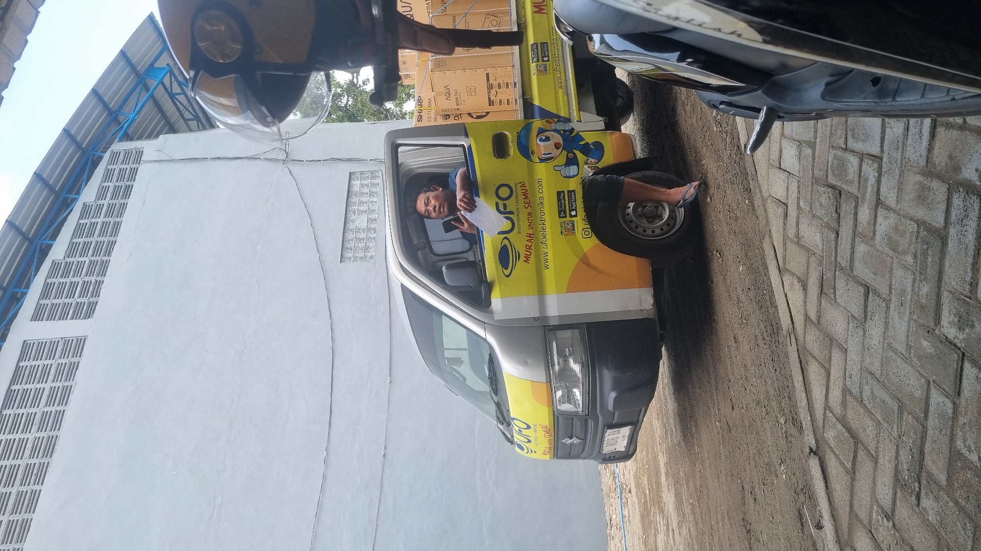 An outdoor, eye-level, medium shot captures a silver and yellow delivery van, branded "UFO ELECTRONIKA," parked on a dirt and paved surface in Diwek, Indonesia. The van is filled with boxed appliances, including brands like Sharp and Aqua. A man is seated in the driver's seat, holding a phone and a piece of paper. He is looking towards the right of the frame. The van's side features a cartoon mascot and the website "www.ufoelektronika.com." In the foreground, the edge of a black car is visible on the right. Behind the van, a white building with horizontal vents and a blue-roofed structure are present. The ground consists of loose dirt and grey interlocking pavers.