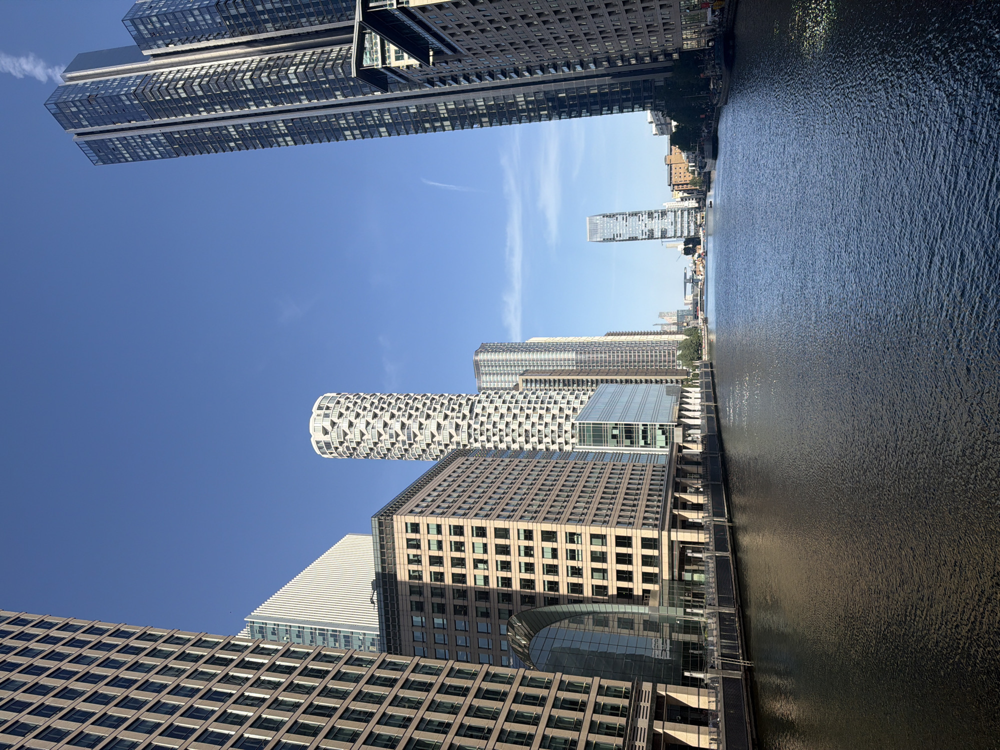 The image shows a low-angle view of the Canary Wharf financial district in London, United Kingdom, as seen from a vantage point along the water's edge. The sky is clear and blue. The river occupies the right side of the frame, and its surface reflects the sunlight. A variety of modern high-rise buildings dominate the skyline on the left, including distinctive architectural styles, such as a tower with a textured, cylindrical design. Other buildings feature glass and steel constructions with rectangular grids of windows. The buildings are aligned along the river, with piers and walkways visible at the water's edge. The image captures the modern urban landscape of Canary Wharf.
