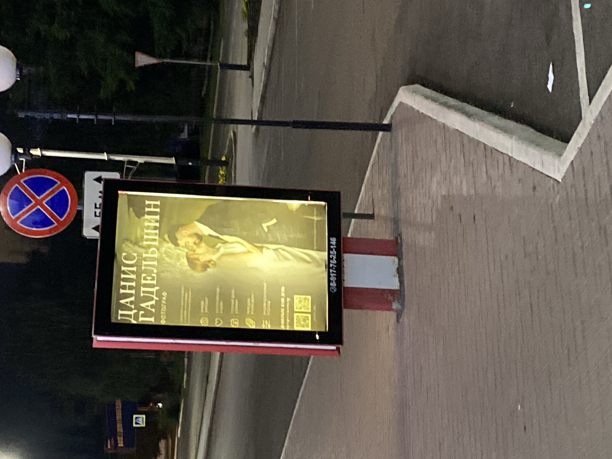 This is a medium shot of a digital billboard in Ishimbay, Russia, located on a sidewalk next to a road. The billboard displays a portrait of a couple, likely for a photography service. The text on the billboard, in Russian, includes "ДАНИС ГАДЕЛЬШИН" (Danis Gadelshin) and "ФОТОГРАФ" (Photographer). Below the image are icons and text indicating services offered, such as "СВАДЕБНЫЕ ФОТО" (Wedding Photos), "ФОТОСЕССИИ" (Photoshoots), and "ПОРТРЕТЫ" (Portraits). At the bottom of the billboard is a phone number, "8-917-76-25-146", and two QR codes. The billboard is mounted on a red and white striped pole. In the background, a street sign with a blue circle and a red X is visible, indicating no stopping or parking. Trees and streetlights are also present in the background, suggesting an urban environment at dusk or night. The sidewalk is paved with brick, and the road is asphalt.