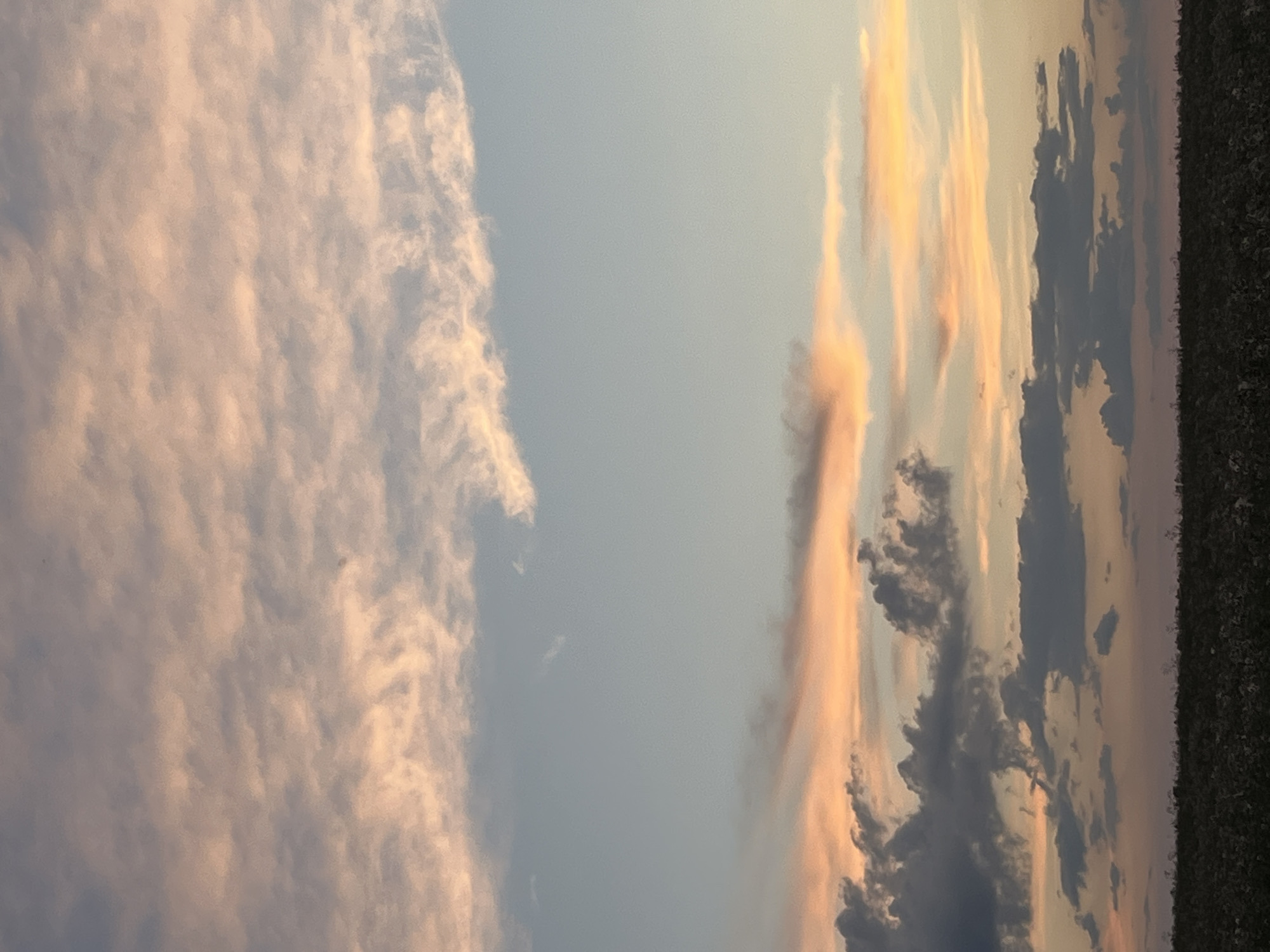 This is a photograph of a sunset over a field in Малая, Russia. The sky is filled with clouds, some of which are illuminated by the setting sun, casting a warm orange and pink glow. The clouds are of various shapes and sizes, with some appearing wispy and thin, while others are more dense and dark. Below the sky, a dark field stretches across the bottom of the image, appearing to be a crop field. The overall scene is one of natural beauty and tranquility, typical of a rural sunset.