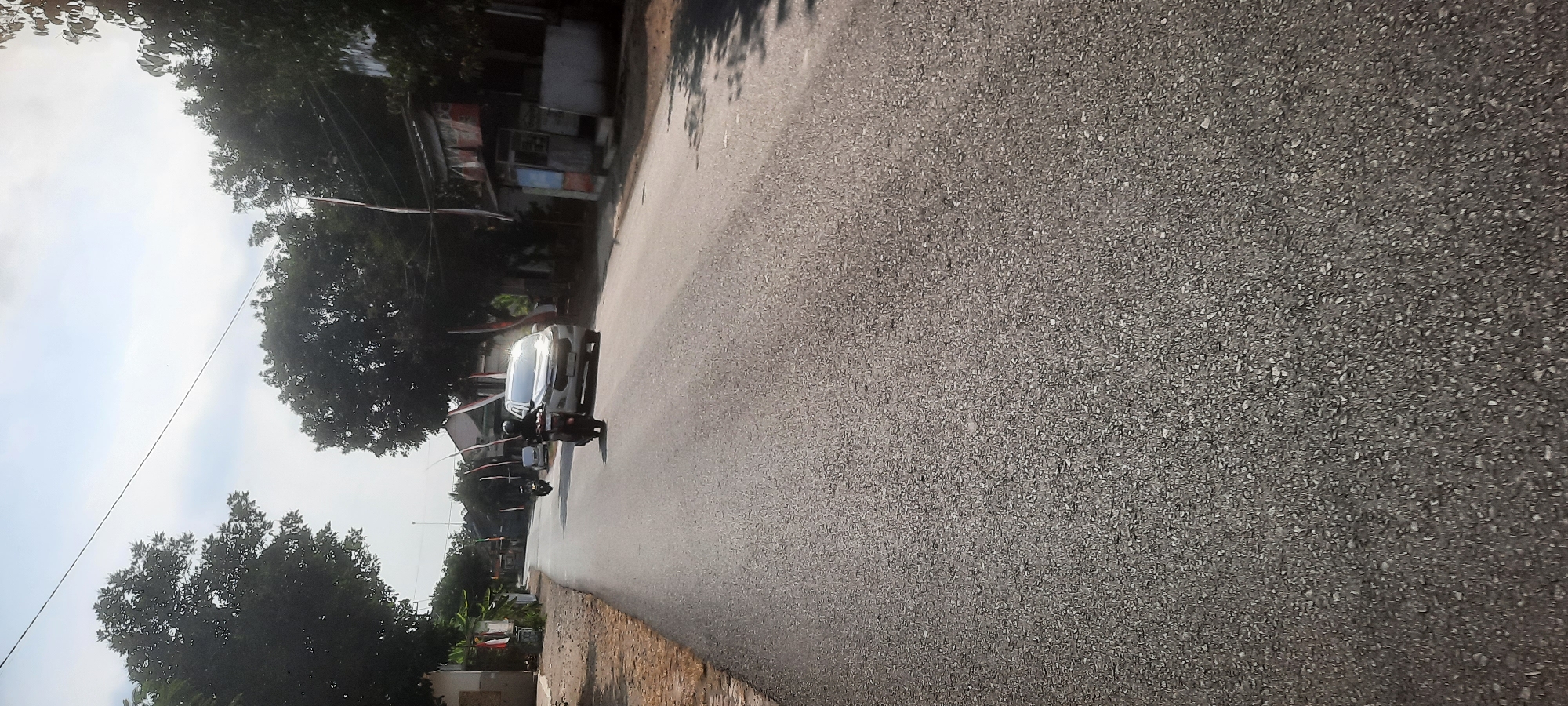 A motorcycle rider is traveling down a paved road in Siraman, Indonesia. A silver SUV is following closely behind the motorcycle. Both vehicles are moving away from the viewer. The road is lined with trees and houses. The sky is clear with some clouds. Red and white flags are visible on poles along the side of the road, suggesting a possible national holiday or celebration.