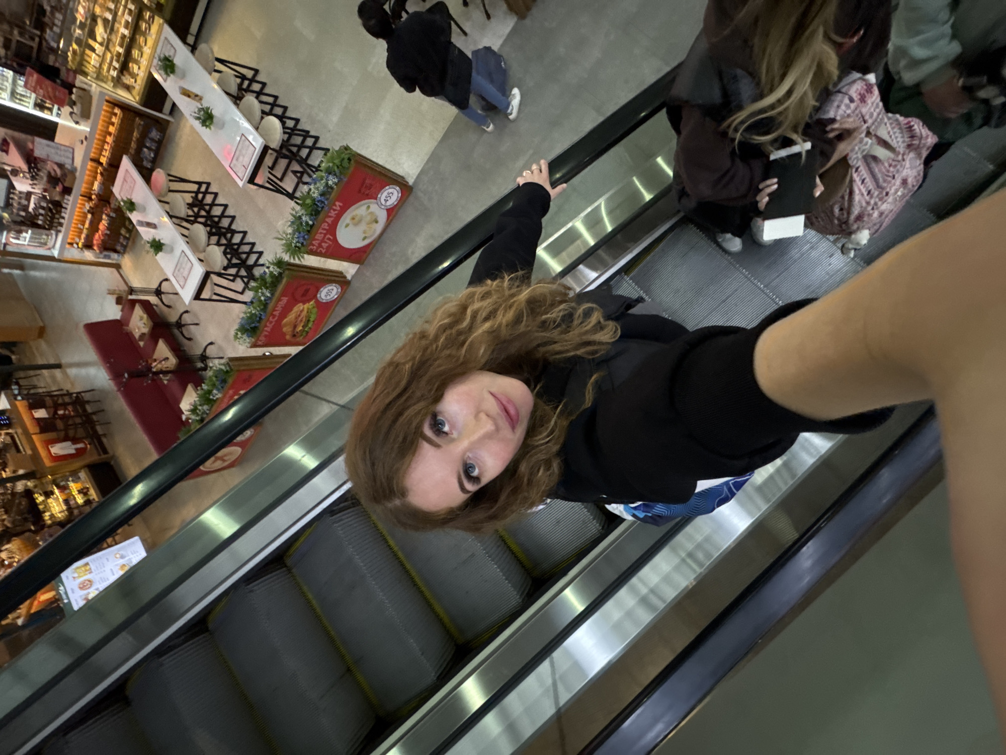 A woman is taking a selfie on an escalator in what appears to be a grocery store or market. The escalator is ascending, with grey steps and yellow accents. The woman has long, wavy red hair and is wearing a black jacket. She is holding onto the escalator handrail with her left hand. In the background, there are other people on the escalator and in the store, including a person in a black jacket and jeans. Store displays feature images of food. The scene is brightly lit. There is no evidence to specifically confirm that the location is the city of Zarechnaya (Gamovskoe S/P), Russia.