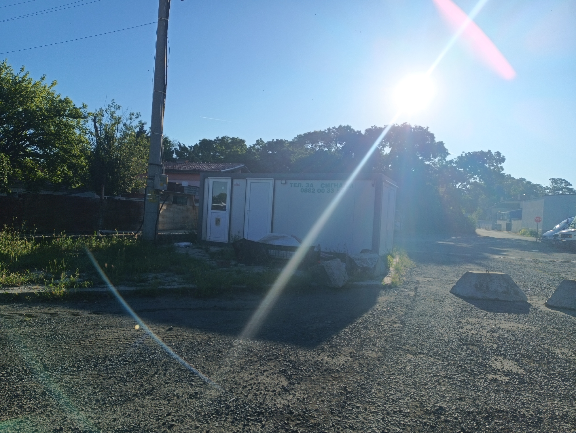 The image depicts a street scene in Karnobat, Bulgaria, under a bright, sunny sky with lens flare prominently visible. In the foreground, a gravel road leads towards a white portable building or container. This building has two doors and what appears to be signage in a foreign language, possibly Bulgarian, with the text "ТЕЛ. ЗА СИГНАЛ" and a phone number "0862 00 33". To the left of the building, there is a wooden fence and lush green trees. A utility pole stands to the left of the frame, with electrical wires running across the sky. In the background, more trees and buildings are visible, along with a parked car and a stop sign. The overall impression is one of a semi-urban or industrial area on a clear day.