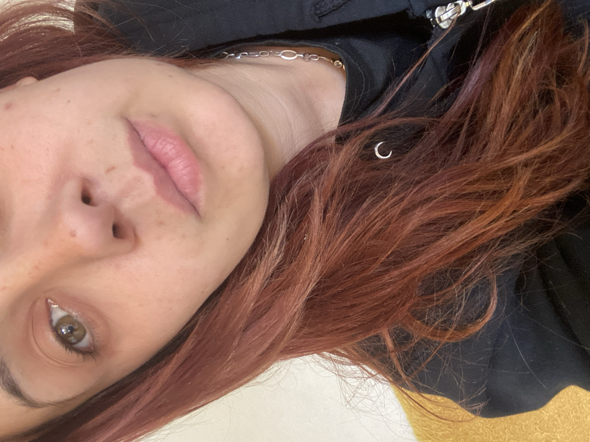 A close-up, tilted headshot of a fair-skinned individual with freckles, red-brown hair, and hazel eyes. The person is wearing a black jacket and a silver necklace. A small silver hoop earring is visible in the hair. The background includes a light-colored surface and a hint of yellow fabric. The location is not discernible from the image and is not in Getsovo, Bulgaria.