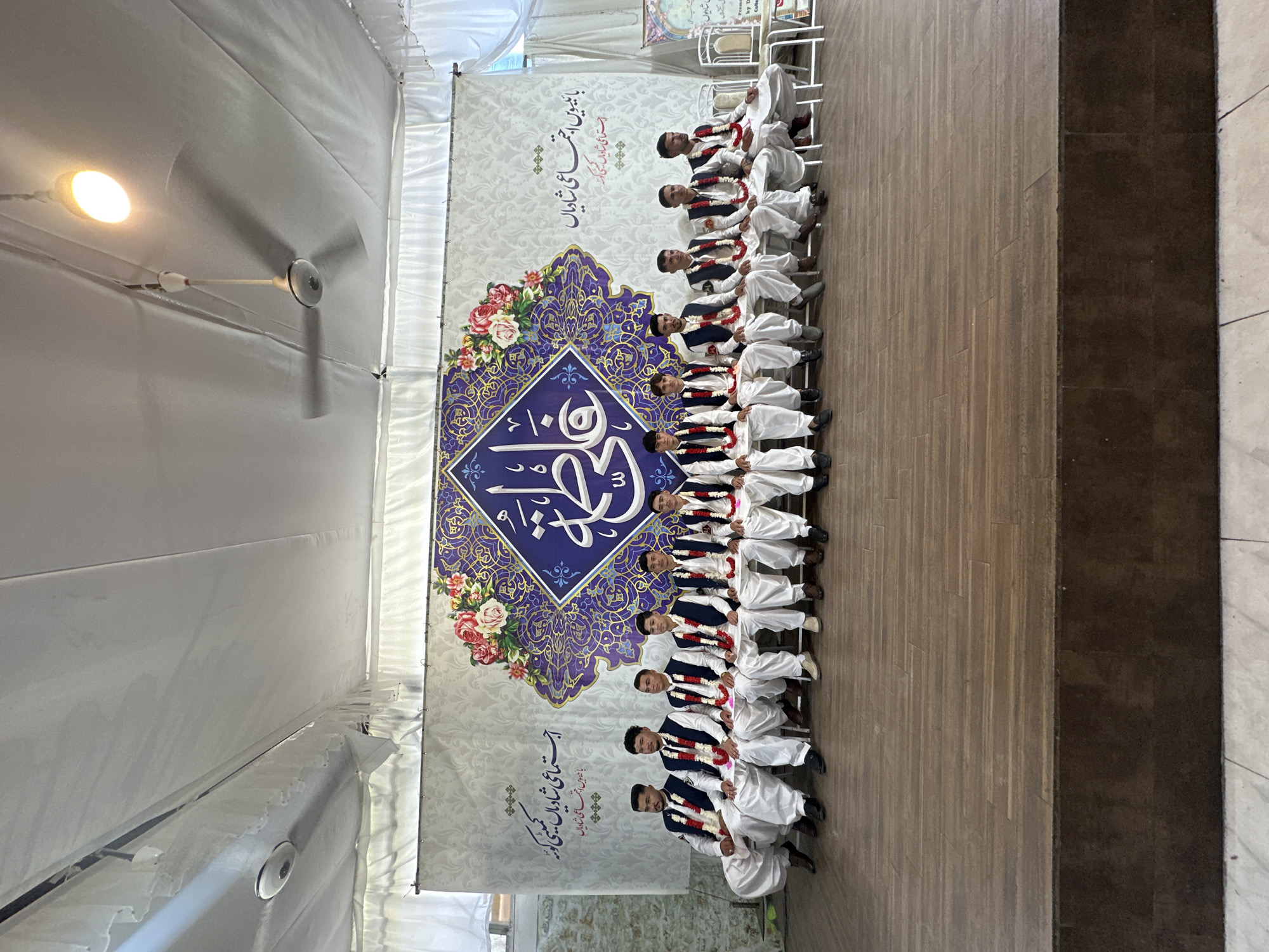 **Scene:** An indoor event in Quetta City Tehsil, Pakistan. A group of men wearing traditional white clothing and dark vests with garlands are seated in a row, likely for a celebration.
**People:** The men are the central subjects of the image. They are uniformly dressed, suggesting a group activity or performance.
**Objects:** A large banner with Arabic text and floral designs is behind the men. There are folding chairs for seating. The room has a ceiling fan and lighting fixtures. The floor is a mix of wooden and tiled surfaces.
**Actions:** The men are seated, and they are likely participating in or posing for an event.
**Interactions:** The men's close seating arrangement suggests they are part of a collective activity.
**Environment:** The event is taking place in a tent-like structure with fabric walls, possibly a temporary setup for a special occasion. The location is in Quetta City Tehsil, Pakistan, based on the text on the banner.