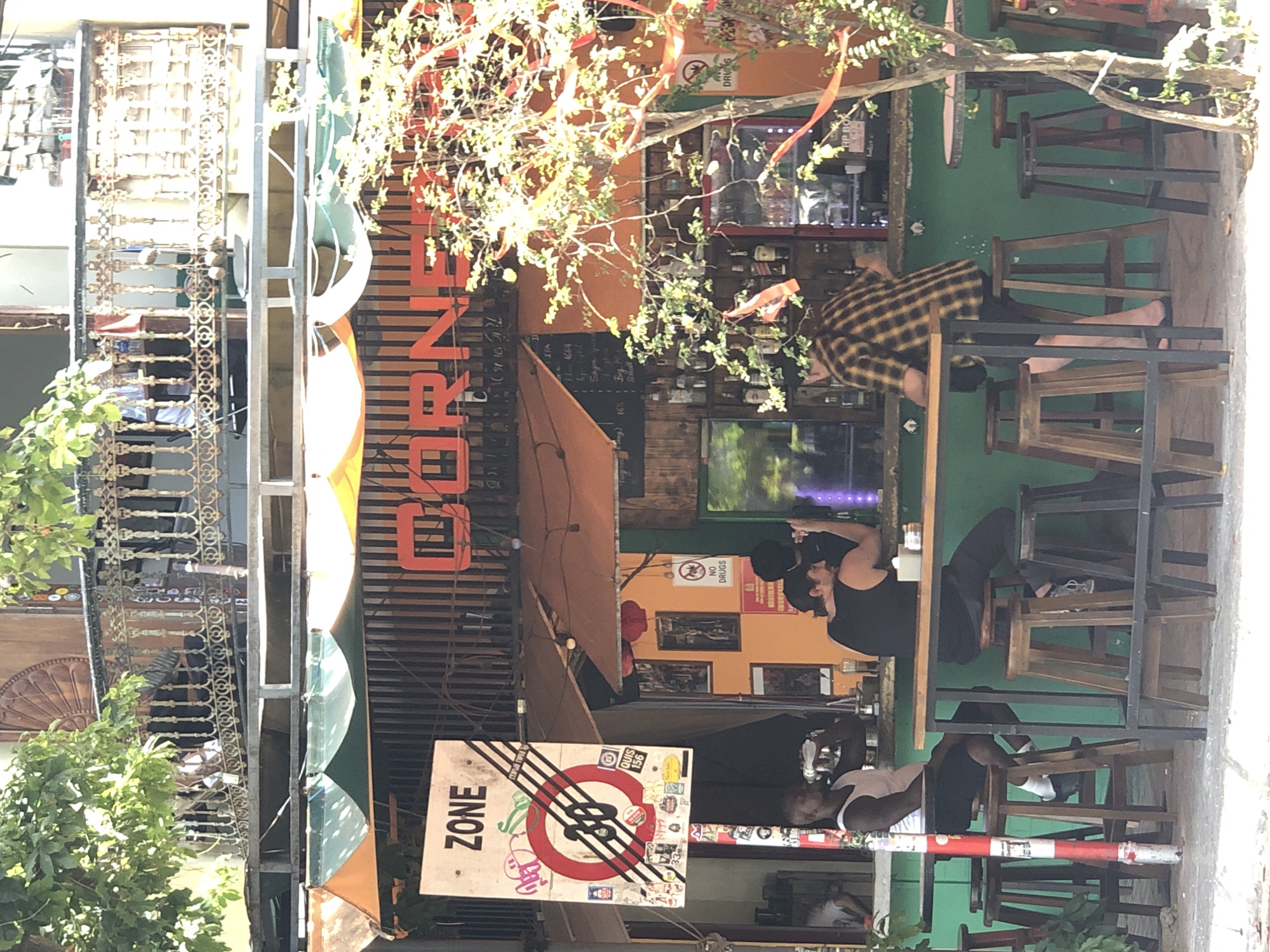 The image shows an outdoor seating area of a bar or cafe in Đà Nẵng, Vietnam. The establishment appears to be named "CORNER" as indicated by the prominent red lettering above the counter. The bar is set against a bright green wall, with shelves stocked with bottles behind it. An orange awning extends over the counter area.
In the foreground, several people are seated at a high bar table. On the left, a man with dark skin and a white tank top is drinking from a bottle while sitting on a wooden stool. To his right, a man with dark hair and a black tank top is seated with his back partially to the camera, facing the bar. Further to the right, a person wearing a yellow and black plaid shirt and black shorts is also seated at the bar, with their back to the camera.
Various objects are visible, including stools, tables, bottles of alcohol on the shelves, a sign with the word "ZONE" and a circular symbol with "30" inside, suggesting a speed limit. The area is decorated with red ribbons tied to tree branches and string lights. The overall ambiance suggests a casual, lively atmosphere typical of a street-side establishment. The architecture of the building behind the bar, particularly the ornate balcony, hints at a blend of colonial and modern styles.