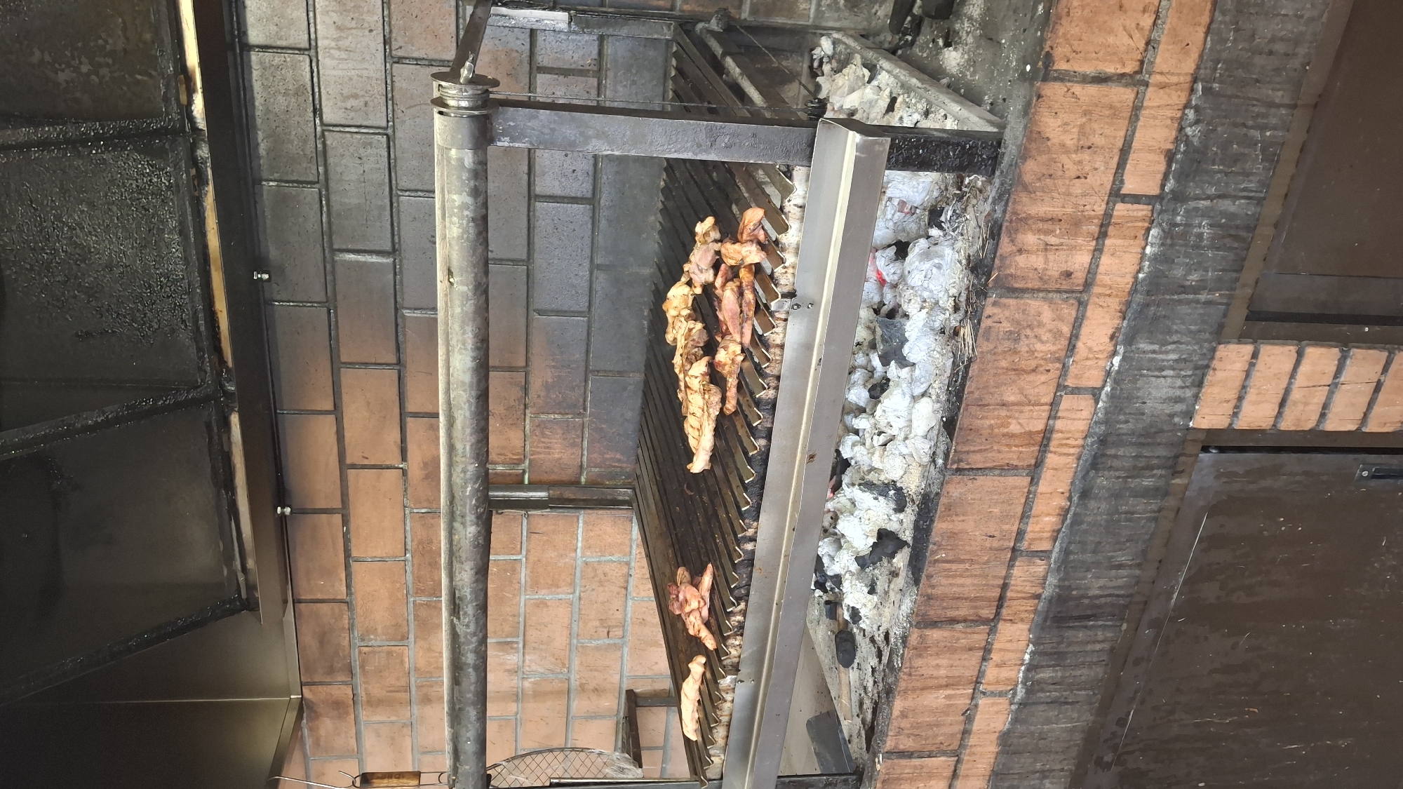 The image shows a large, outdoor brick barbecue grill with meat cooking on the grate. The grill has a metal frame and is filled with hot coals. The background consists of a brick wall and a stainless steel hood. The overall scene suggests a culinary setting, possibly in a restaurant or a dedicated outdoor cooking area in La Puebla de Híjar, Spain.
