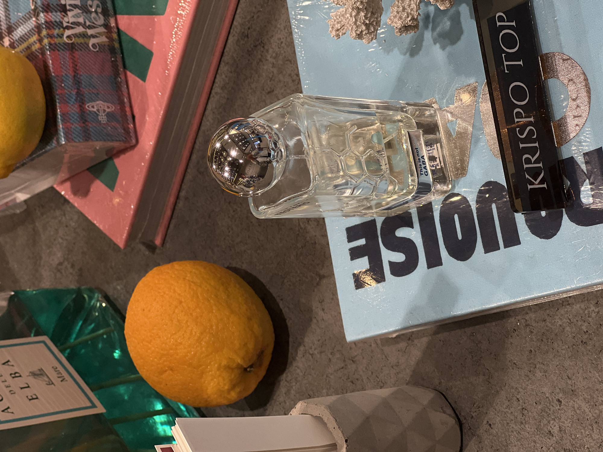 The image depicts a still life arrangement on a dark, textured countertop. Dominating the foreground is a clear glass perfume bottle with a silver, faceted spherical cap. The bottle contains a pale yellow liquid and has a honeycomb pattern embossed on its sides. A small label on the bottle reads "This is not GABA".
To the left of the perfume bottle, an orange rests on the countertop. Behind the orange is a tall, rectangular translucent teal bottle with a label that says "ACQUA DELLA ELBA Mare".
In the background, a stack of books is visible. The top book is pink and has a small box with a tartan pattern and a Vivienne Westwood logo on it. A yellow lemon is partially visible on top of this box.
Beneath the perfume bottle and to its right is a light blue surface, possibly a book or mat, with dark lettering on it, partially obscured. Part of the lettering reads "NOISE" and below that, a black rectangular plaque with "KRISPO TOP" printed in white text is placed on the surface. To the right of the perfume bottle and the blue surface, a piece of white, textured coral is visible.
The lighting suggests an indoor setting with overhead lights reflecting off the shiny surfaces. There are no people visible in the image, and no specific landmarks or details indicate a location in Klaipėda, Lithuania.