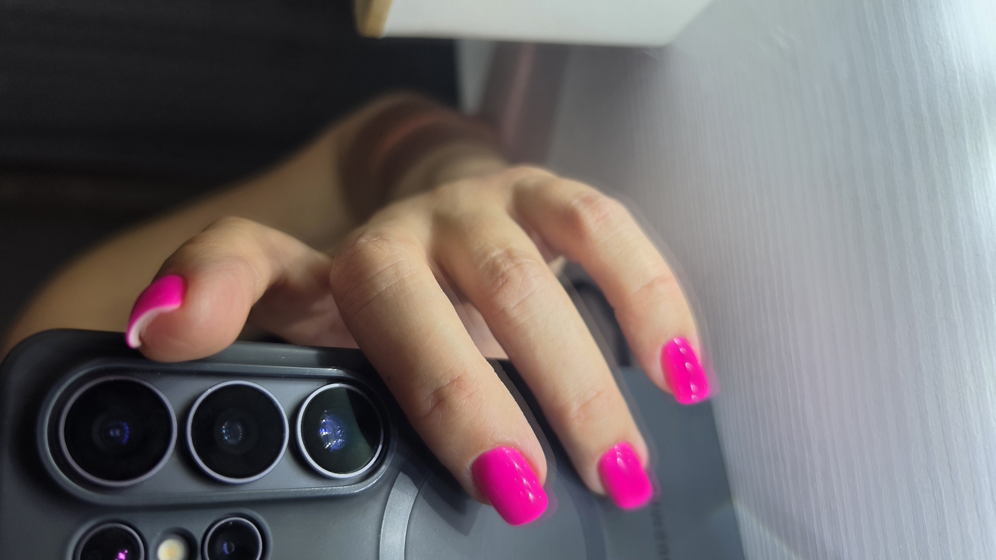The image shows a close-up of a person's hand with bright pink fingernails, holding a dark grey smartphone with a camera array. The hand is positioned on a white, textured surface. The background is dark and out of focus.