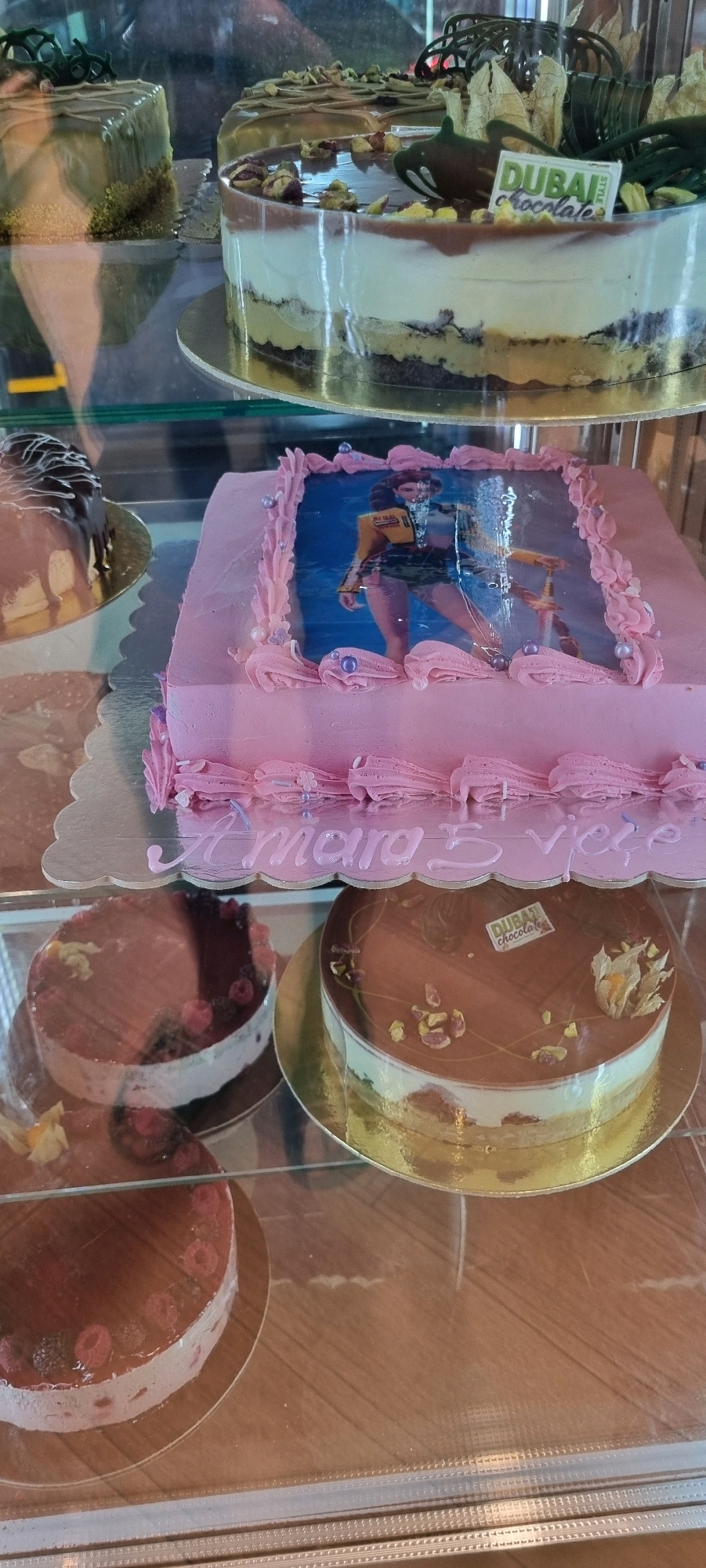 This is a close-up, eye-level, vertical shot of cakes displayed in a glass case, likely a bakery or patisserie. The lighting is natural, coming from above and slightly to the left, creating reflections on the glass.
In the foreground, a rectangular pink cake is adorned with piped frosting flowers and pearls. An edible image of a woman in a yellow jacket and shorts, holding a sword, is centered on top. Below the cake, "Amara S. Veçe" is written in white icing.
To the right of the pink cake, a round chocolate cake with white cream layers and a chocolate ganache topping is visible. It's decorated with pistachios and a small orange fruit. A "Dubai Chocolate" label is placed on top.
Below these cakes, two more round cakes are partially visible. The one on the left is topped with raspberries and dark berries. The one on the right appears to be similar in design to the chocolate cake above, with a similar garnish.
Further back and to the left, a square cake with green frosting and a crumbly base is seen. Another round cake with a dark glaze and decorative elements is also in the background.
The display case is made of glass and metal. The background is out of focus but suggests other baked goods and reflections of the environment. The overall scene is clean and organized, typical of a food service establishment.
This image is located in the city of Bashkia Durrës, Albania.