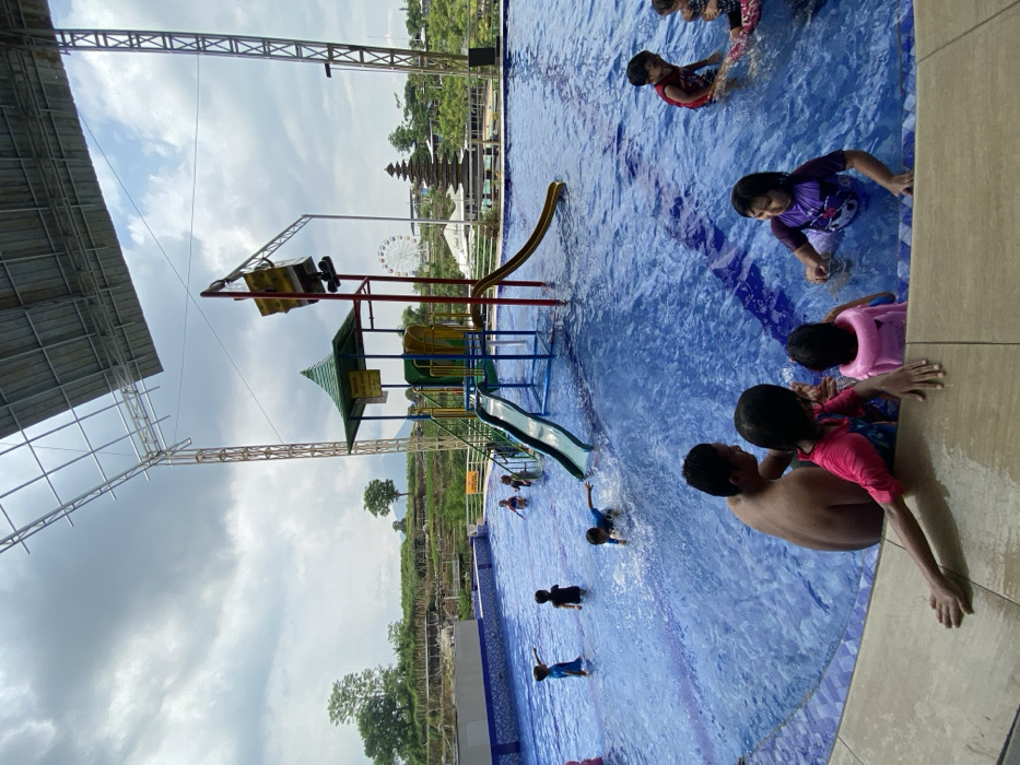 A vibrant outdoor swimming pool with children playing and adults supervising. One area has playground equipment with slides, and in the background there is a Ferris wheel and temple-like structure. The pool is surrounded by green landscape, and overhead is a metal structure. The scene is captured on a partly cloudy day.