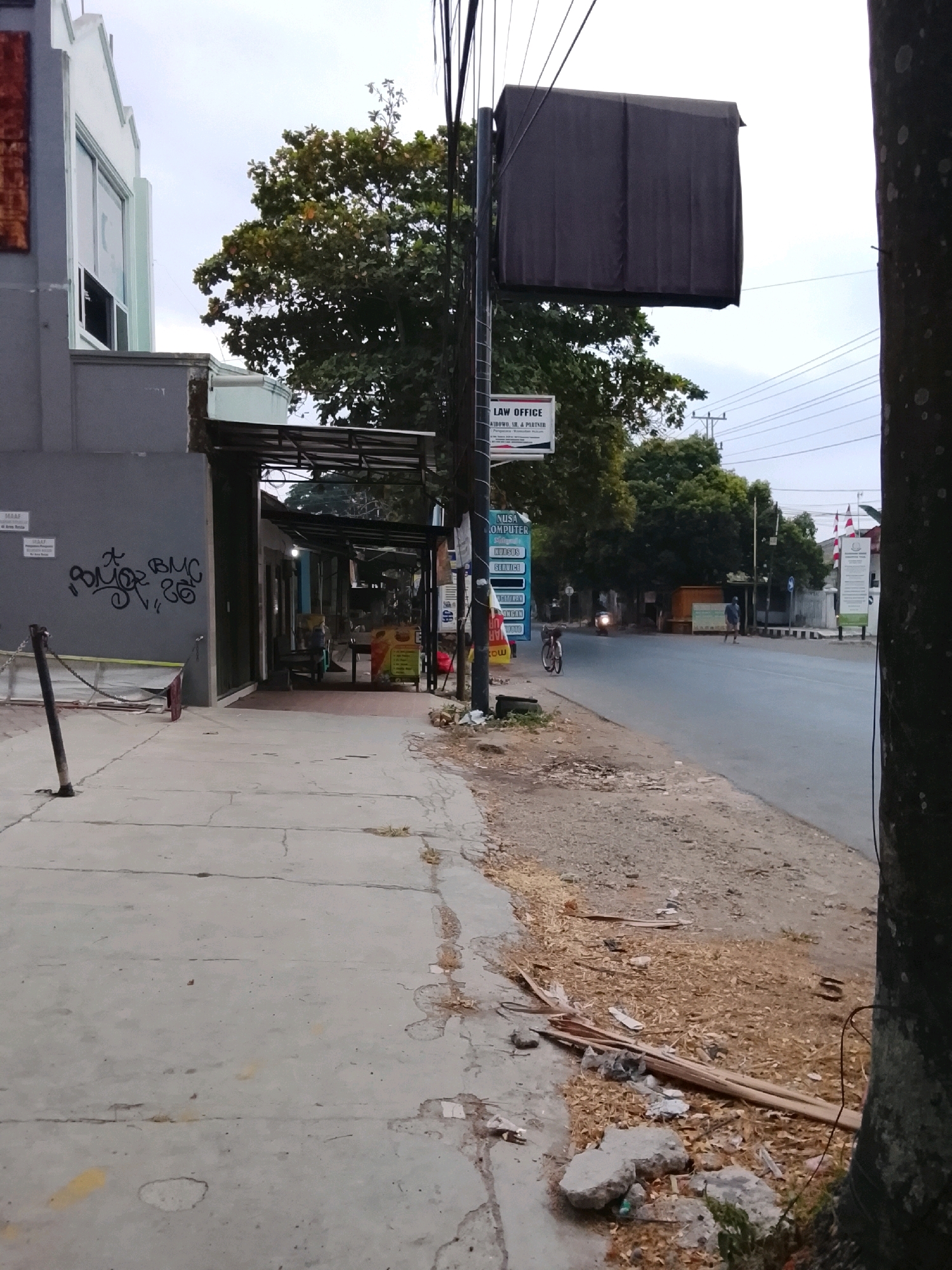 The image depicts a street scene in Slawi, Indonesia, likely in the late afternoon or early evening, judging by the overcast sky and subdued lighting. The foreground is dominated by a concrete sidewalk that appears cracked and weathered in places, with debris such as leaves, twigs, and small stones scattered along its edge. To the left, a gray building with a light-colored upper section and a window is visible. Graffiti in black spray paint adorns the lower portion of this building. A metal post connected to a chain is positioned on the sidewalk.
Adjacent to the building is an open-air establishment with a covered walkway, featuring several stalls or shops. One stall has a colorful sign with text and images. A bicycle is parked near the roadside.
The street itself is asphalt and appears to be a two-way road with moderate traffic. A single motorcycle is seen in the distance, and a person is walking along the side of the road. Overhead, power lines and a dark, fabric-covered sign or screen are suspended from poles. Various other signs, including one for "LAW OFFICE" and another for "NUSA COMPUTER," are mounted on poles along the sidewalk. Trees with green foliage are present, some lining the street and others forming a backdrop. In the distance, more buildings and trees are visible, along with what appear to be Indonesian flags. The overall impression is one of a busy, somewhat commercial urban area.